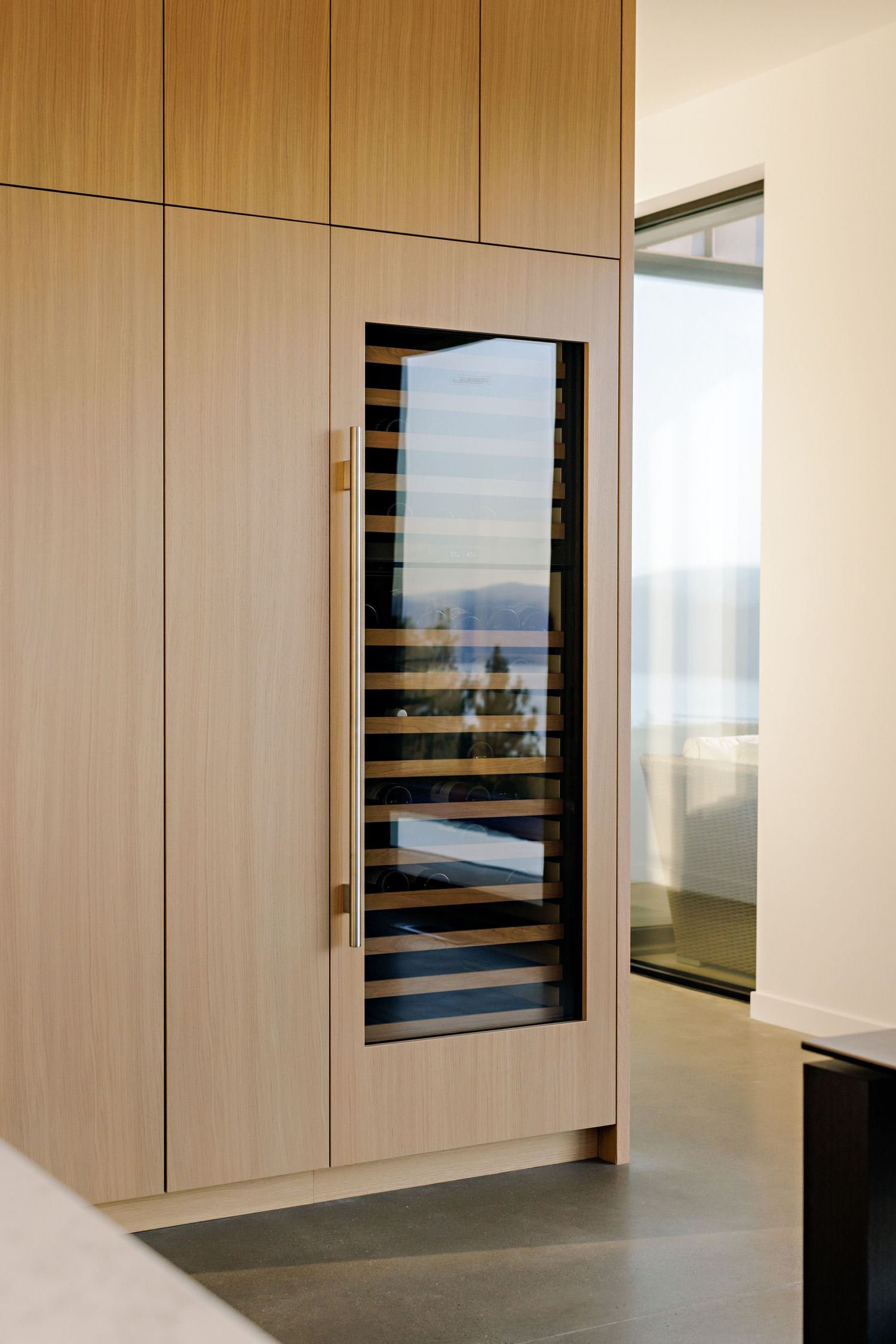 Built-in wine refrigerator with glass door panel mounted in light wood cabinetry showcases rows of stored bottles.