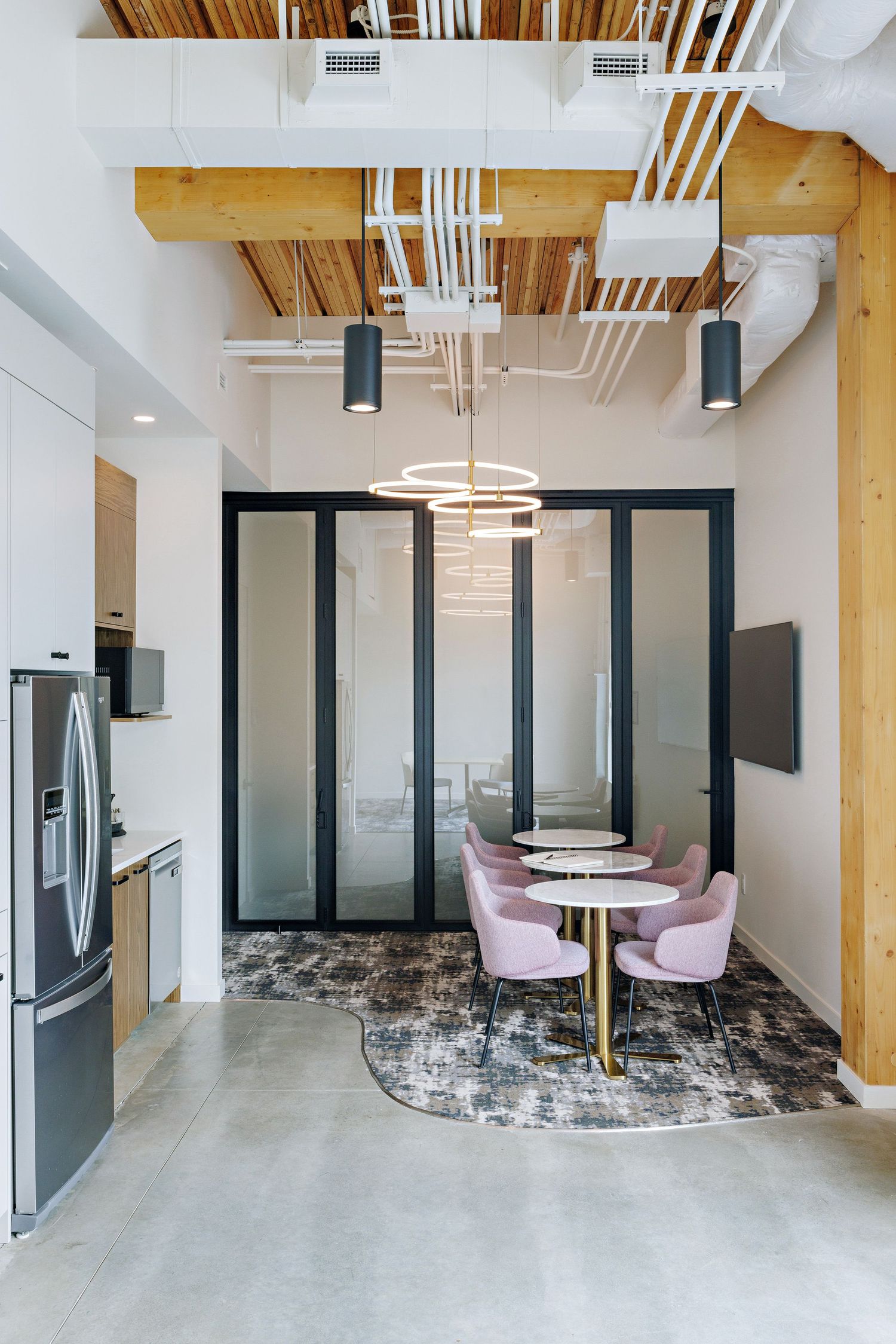 Modern office breakroom with exposed wooden beams, frosted glass doors, pink chairs and circular table on patterned rug.