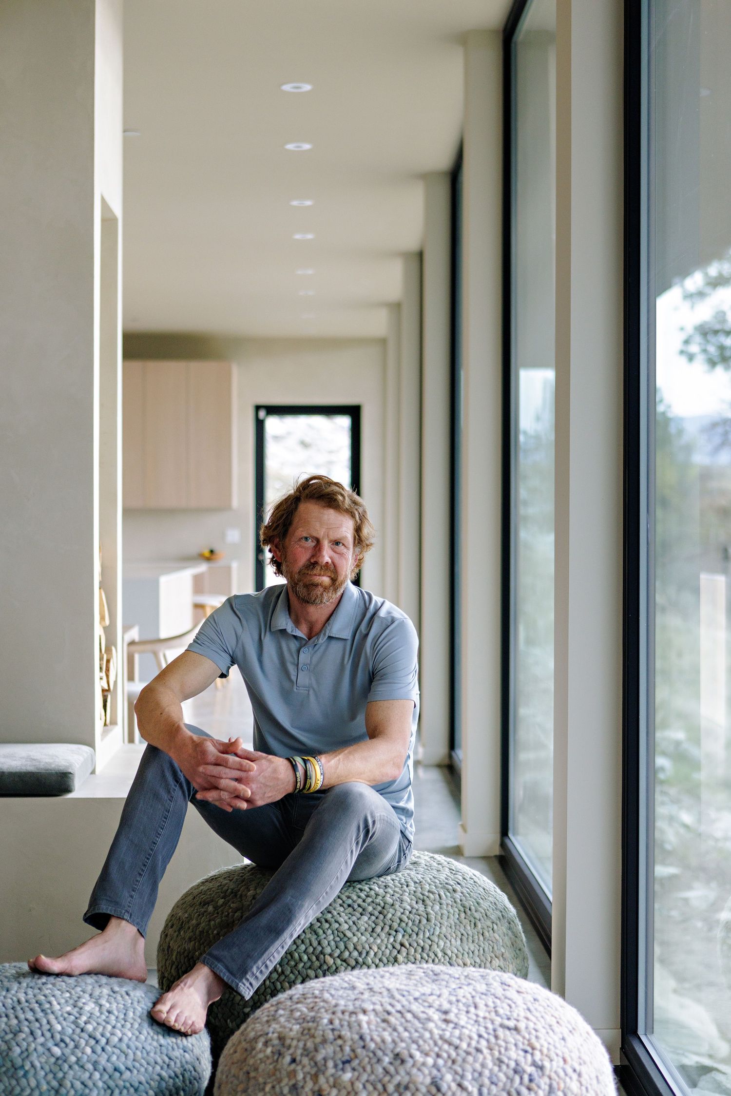 A relaxed person sits on knitted poufs near large windows in a modern, minimalist interior space.