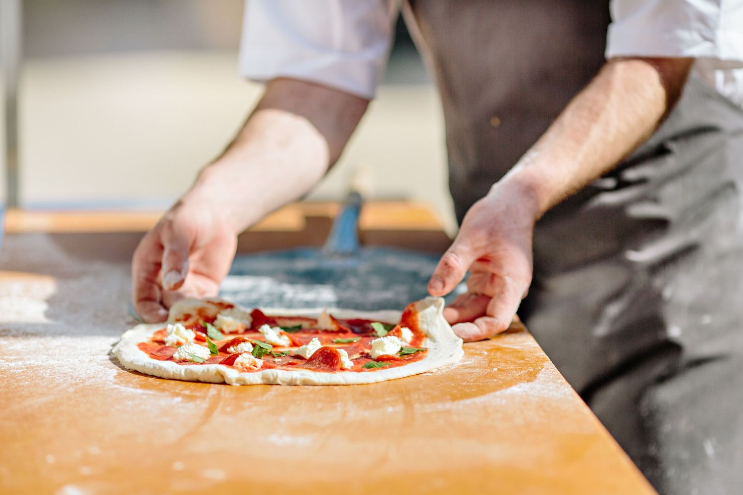 Chef preparing a fresh pizza with toppings on a wooden surface dusted with flour.