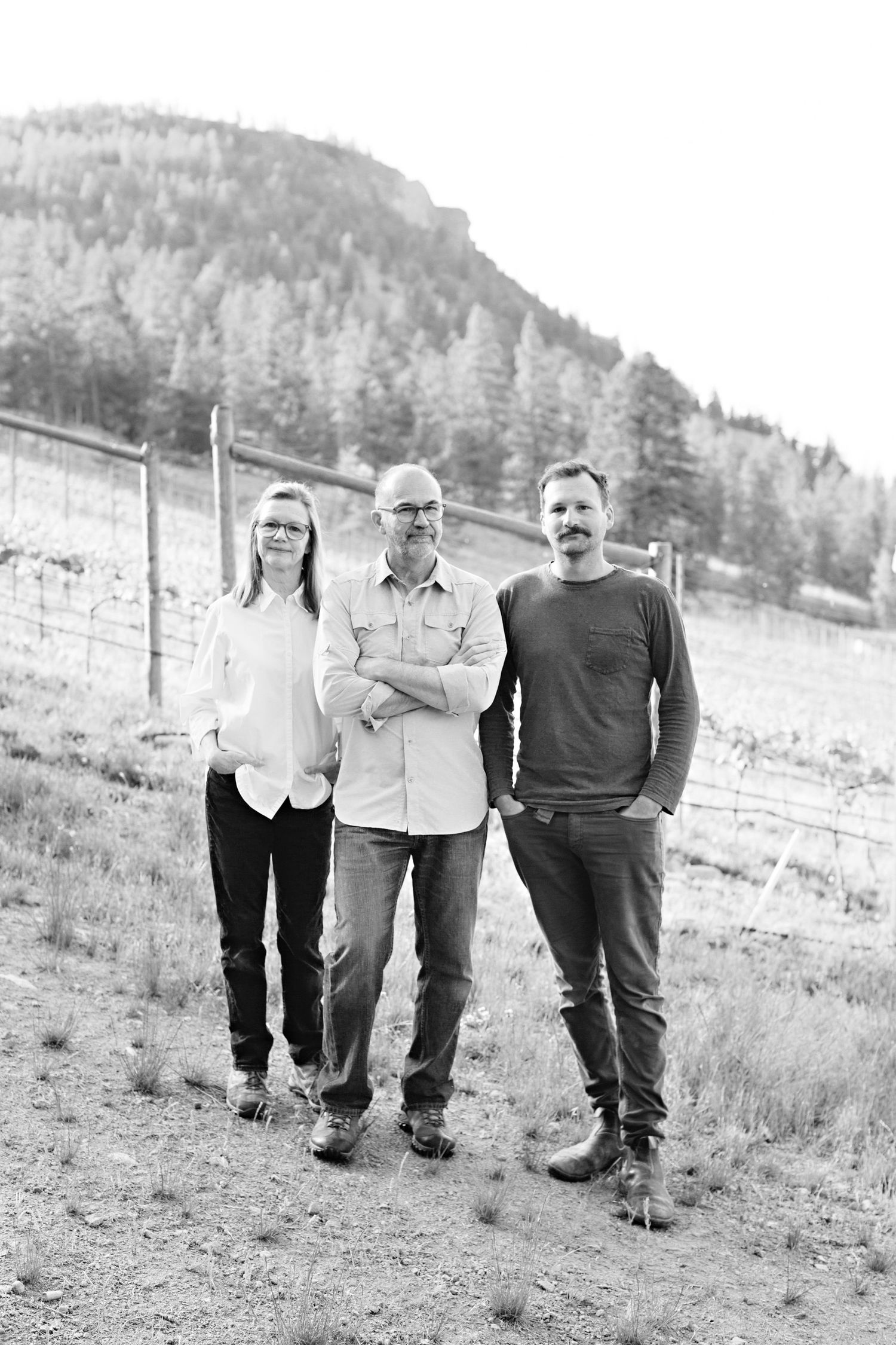 Black and white portrait of three people standing outdoors with a forested hillside in the background.