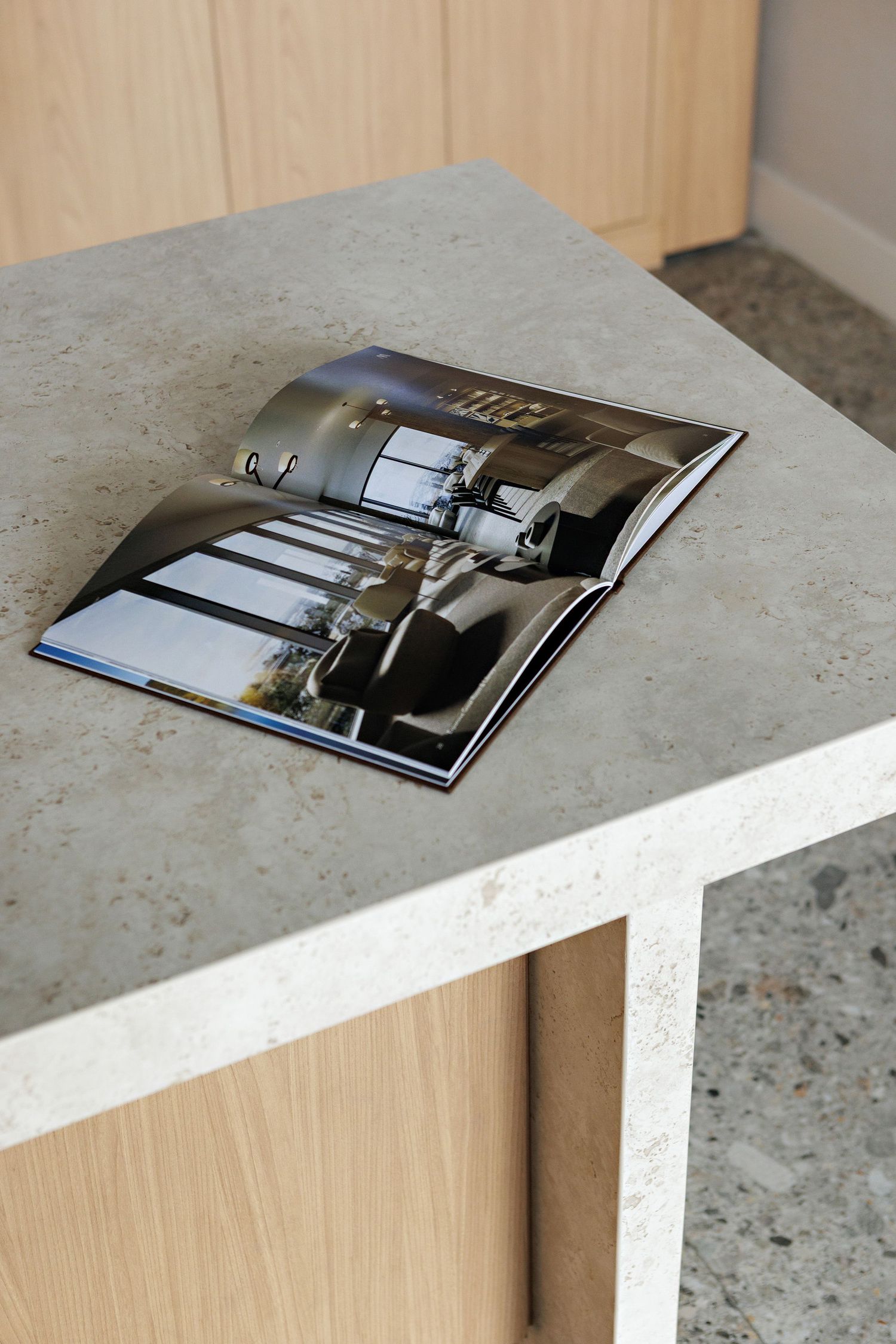 An open magazine displaying interior design photos lies on a white concrete surface near wooden cabinetry.