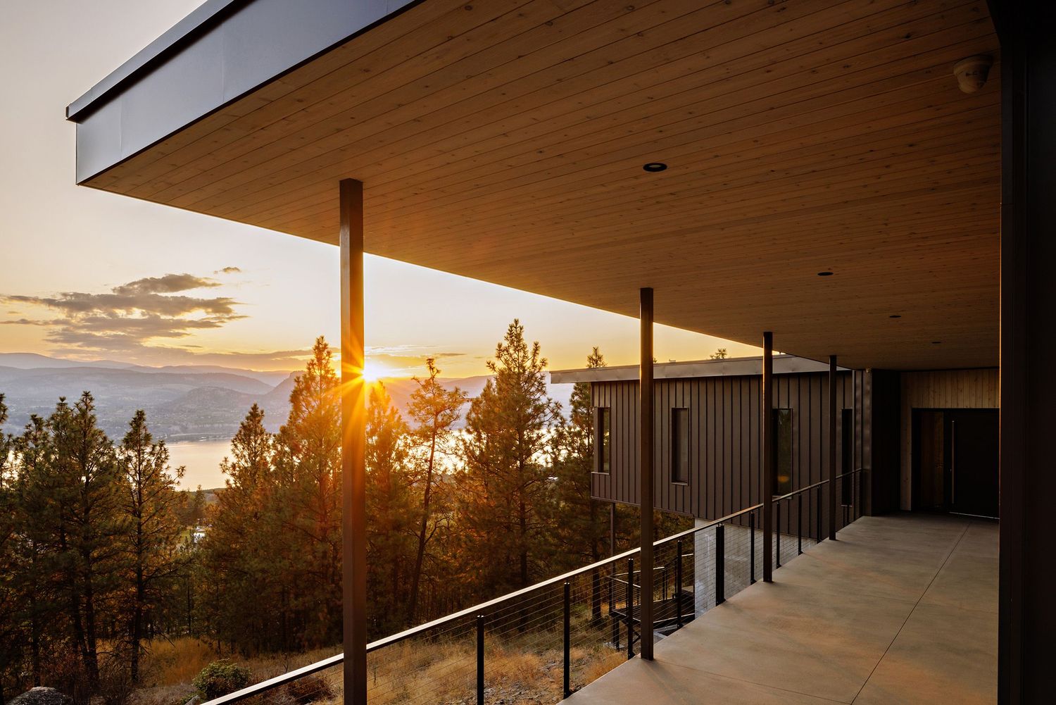 Modern balcony overlooking pine trees and sunset with glass railing and wooden ceiling overhang at luxury residence.