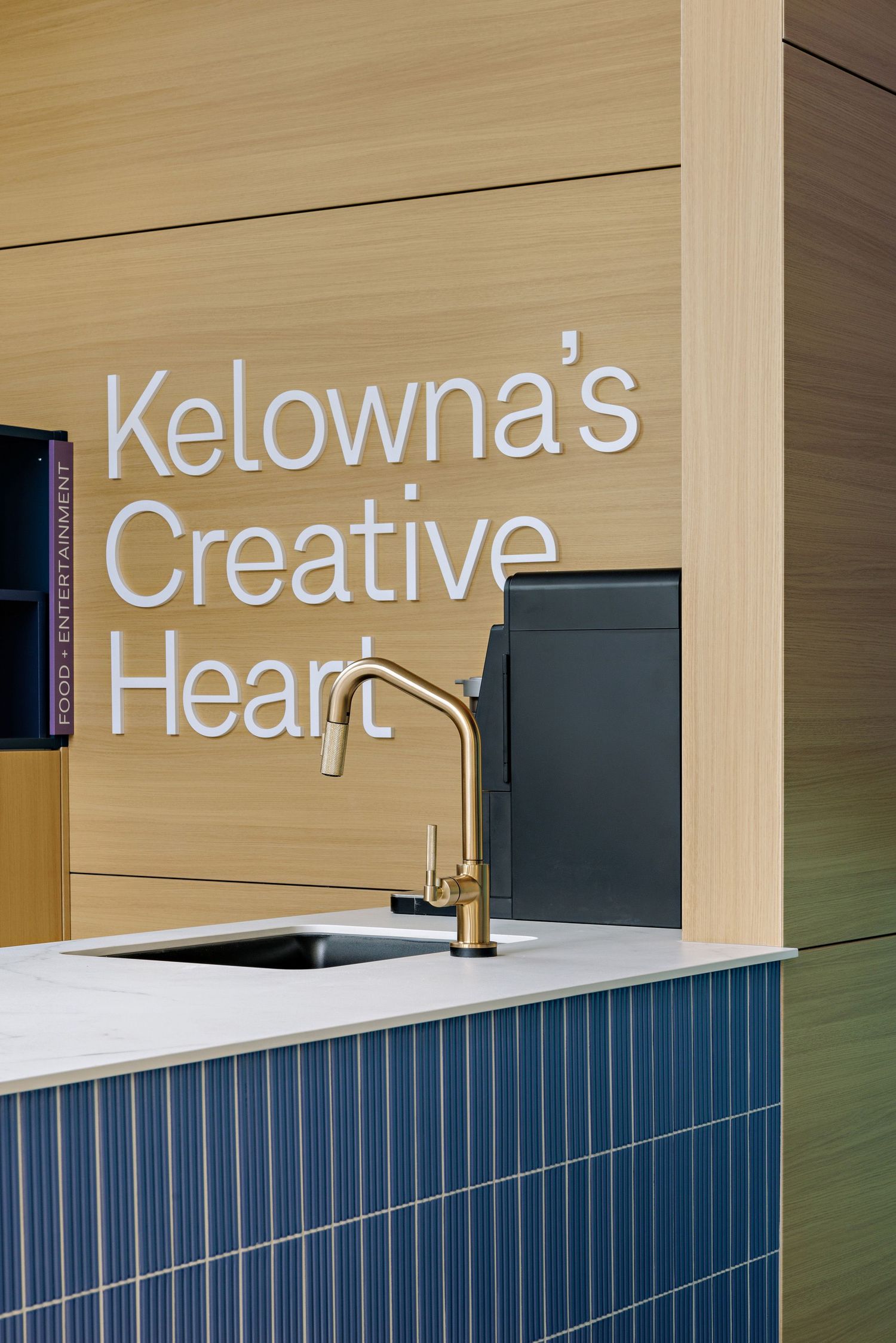 Modern kitchen sink with gold faucet against blue tiled backsplash and wood wall with 'Kelowna's Creative' text signage.