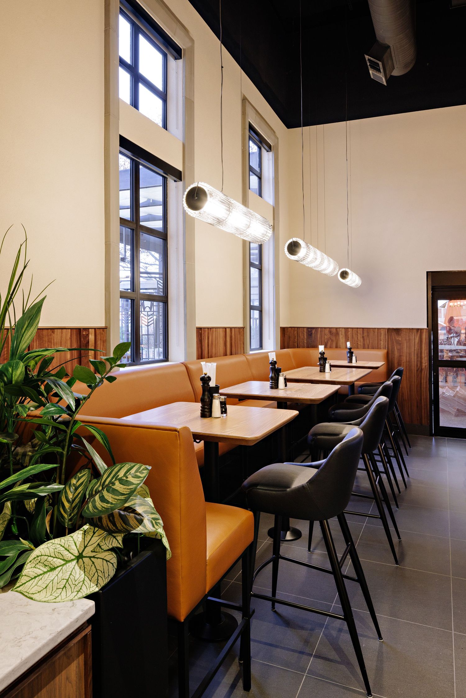 Modern restaurant interior with tall windows, leather banquette seating, black barstools and hanging pendant lights.