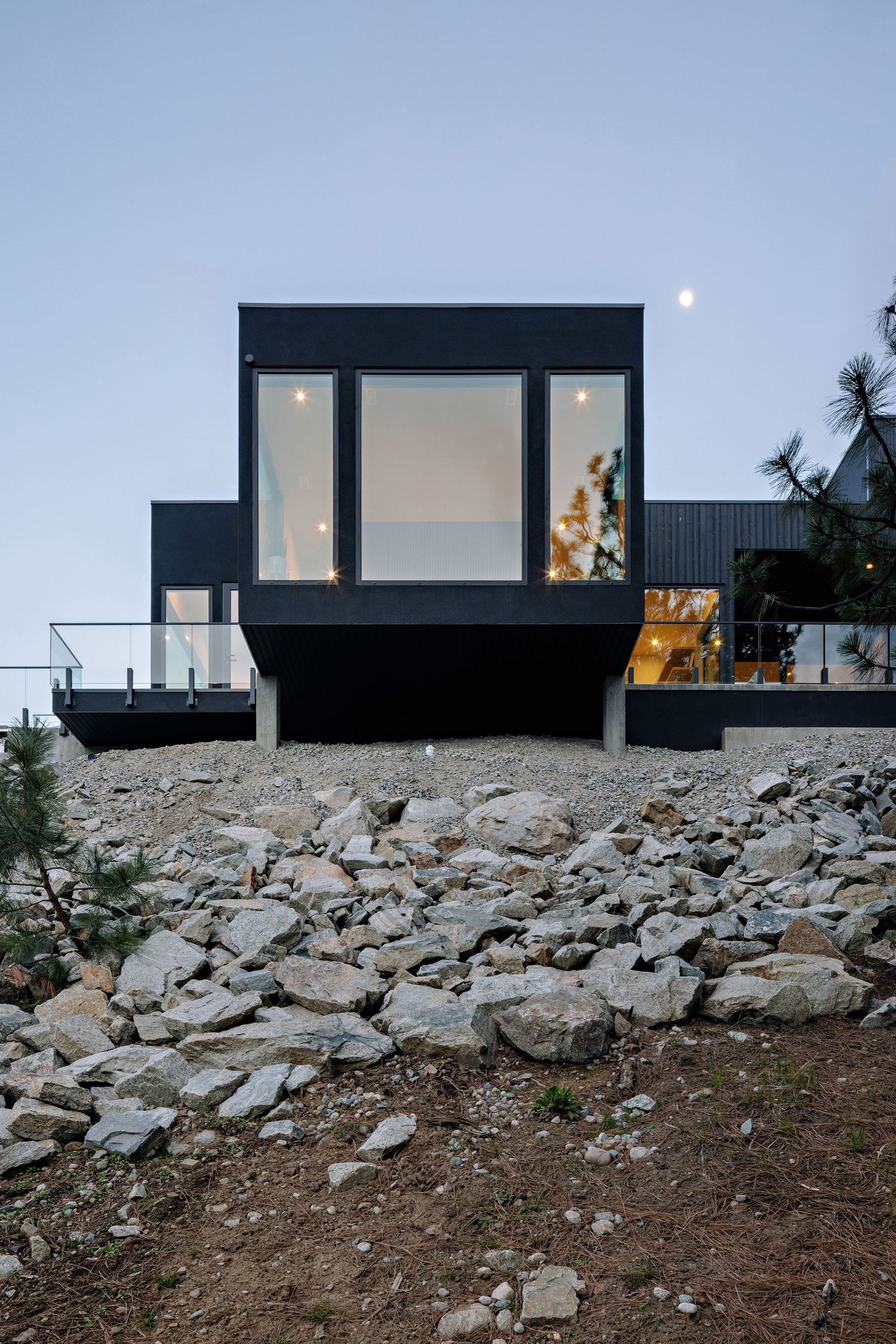 Contemporary black house with large glass windows perches on a rocky hillside at dusk with illuminated interiors.
