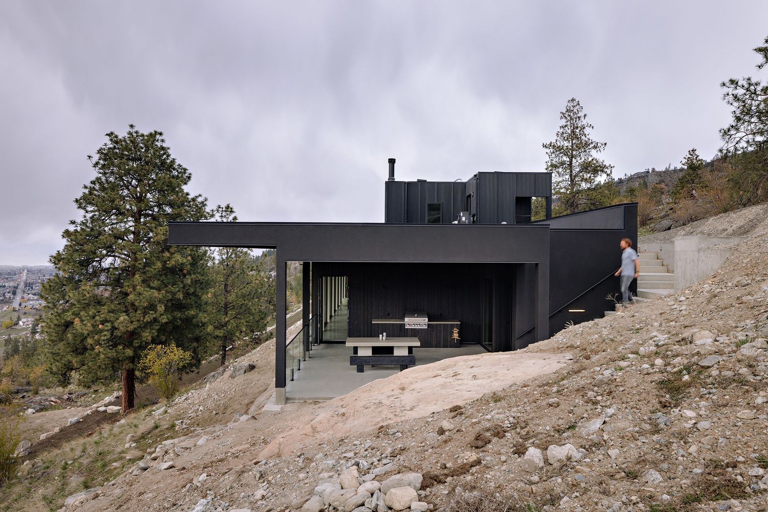 Modern black house with large windows perched on rocky hillside surrounded by pine trees.