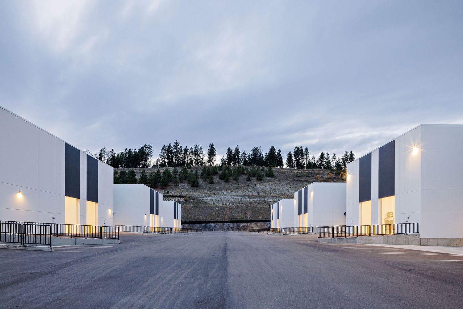 Modern industrial warehouse buildings with white walls and exterior lights line a wide concrete road at dusk.