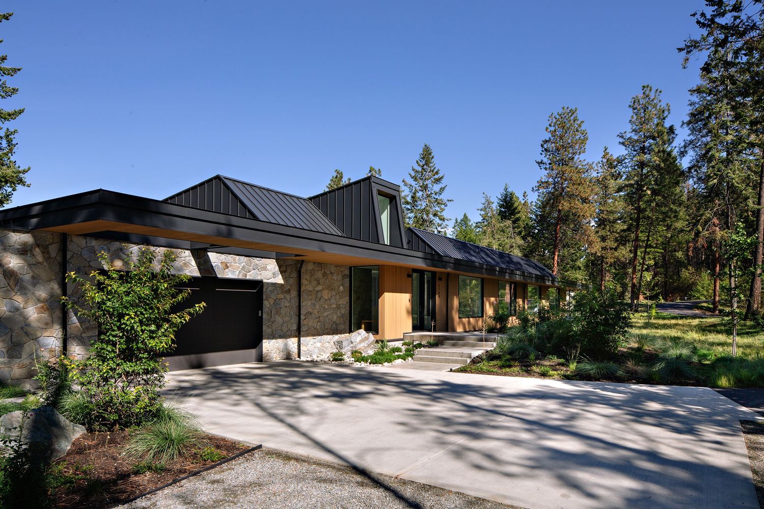 Contemporary ranch-style home with dark roof and stone accents nestled among tall pine trees.