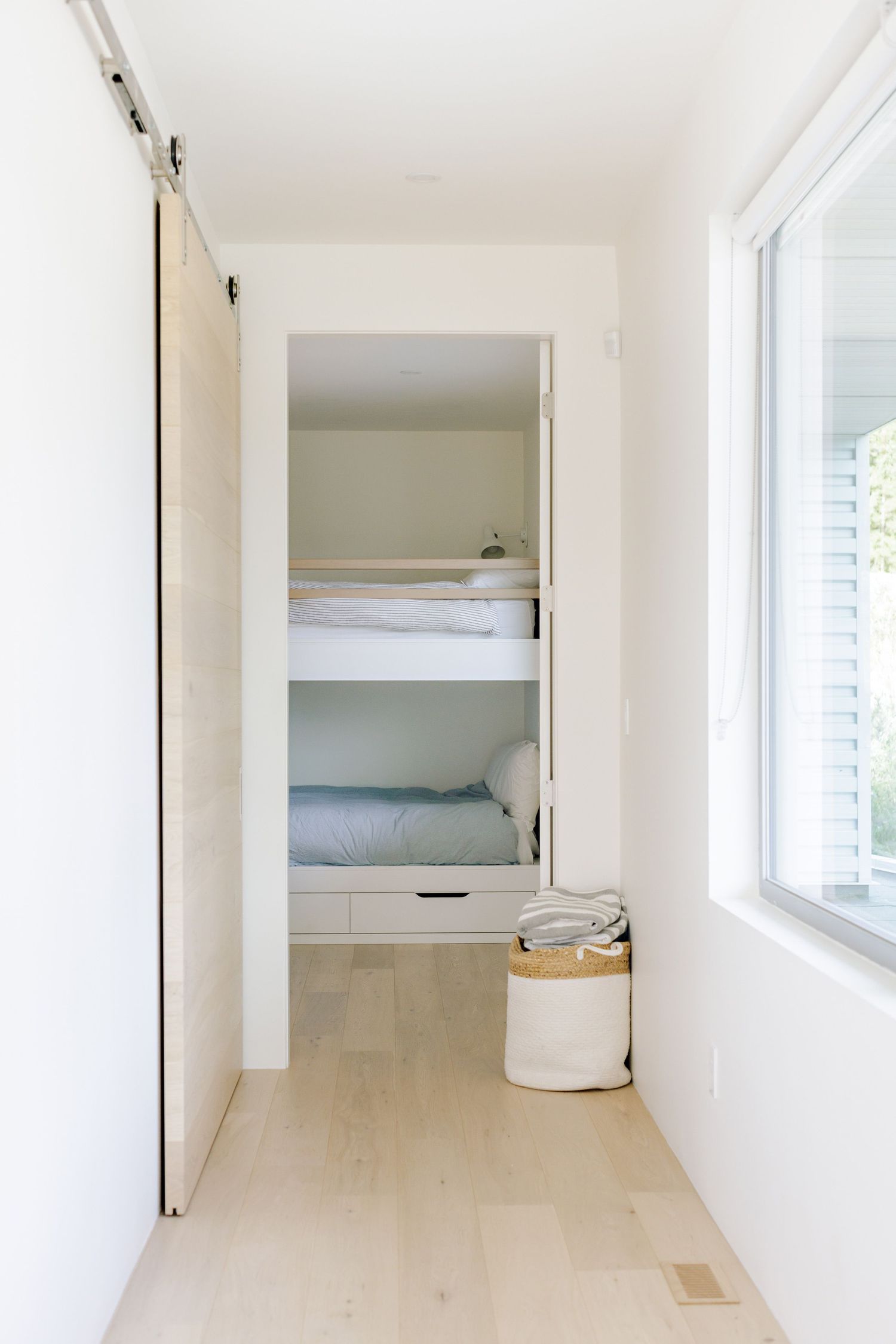 A minimalist white hallway with built-in bunk beds behind a sliding door and a window bringing in natural light.