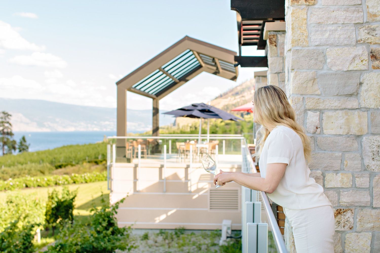 Someone in white enjoys a glass of wine on a sunny terrace overlooking scenic hillside vineyards and mountains.