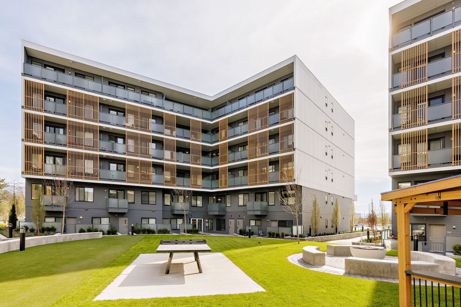 Modern apartment complex with L-shaped design featuring balconies and a landscaped courtyard with picnic tables.