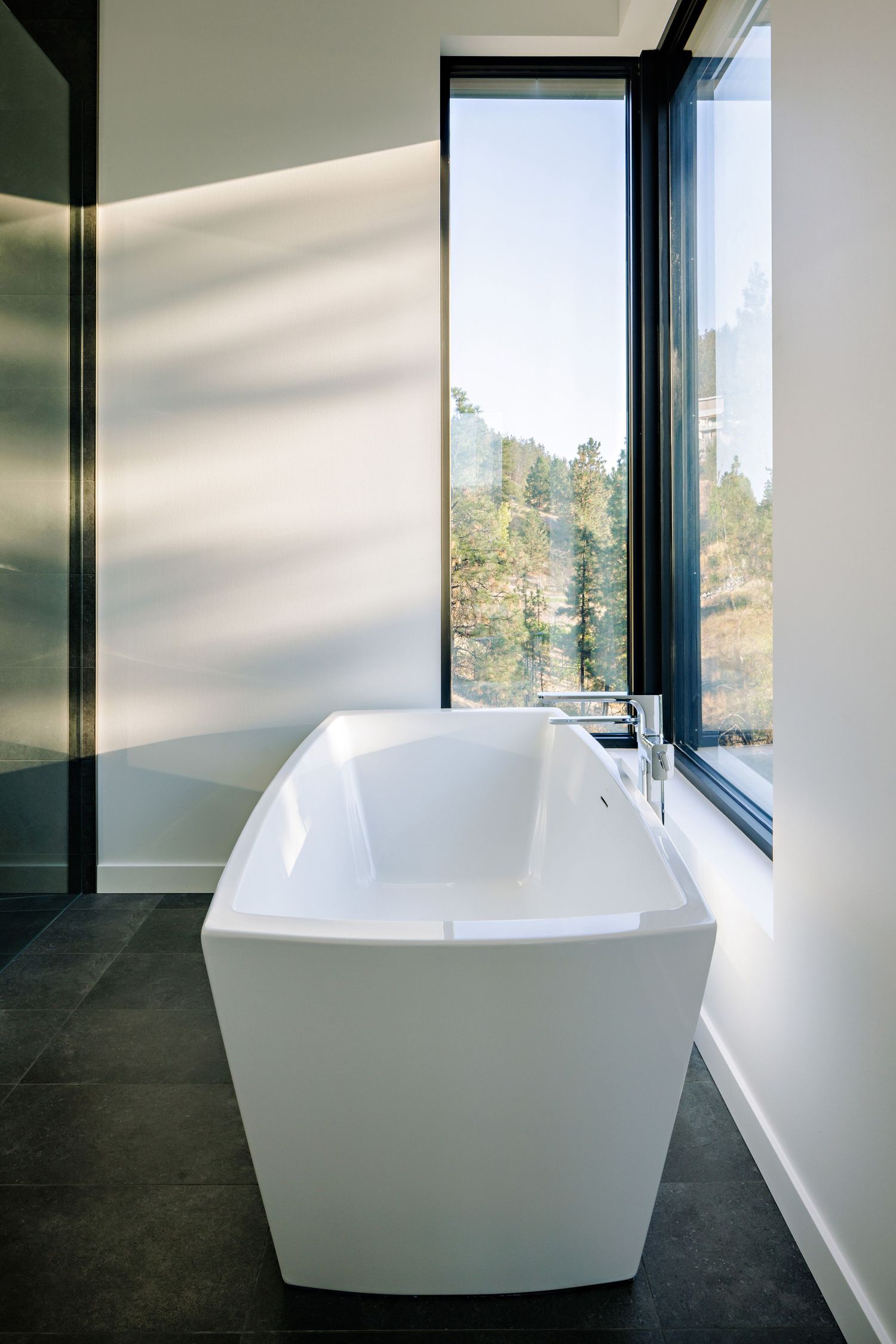 Modern freestanding white bathtub next to floor-to-ceiling windows offering natural light and forest views.