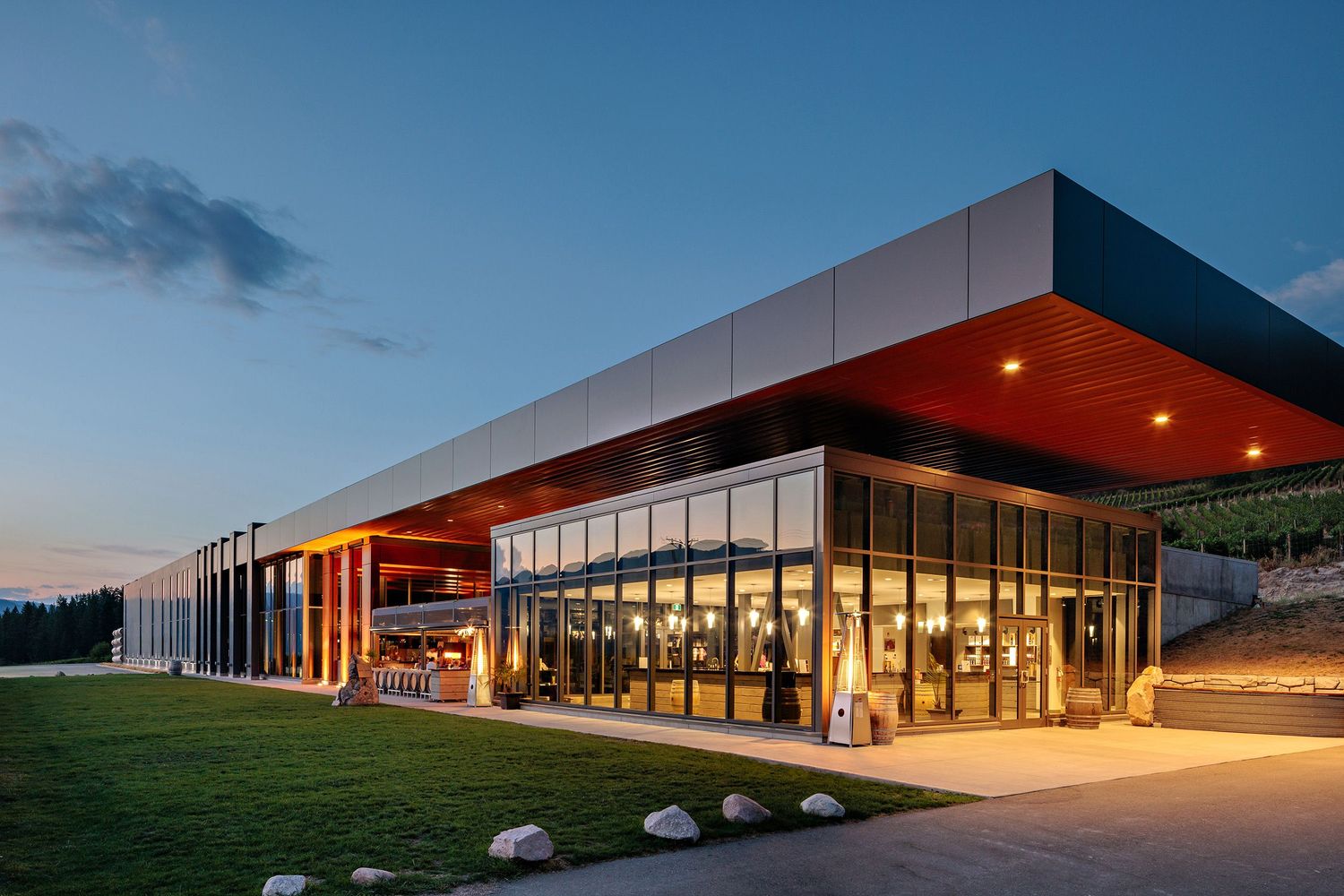 Modern glass and steel building with illuminated interior glows warmly against twilight sky on a green lawn.