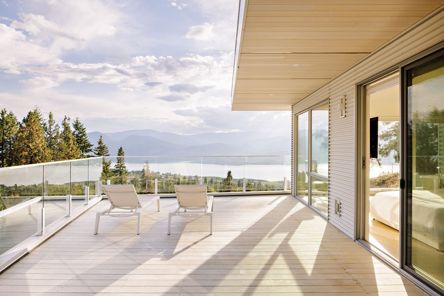 Modern deck with glass railing and lounge chairs overlooking mountain views on a sunny day with sliding glass doors.
