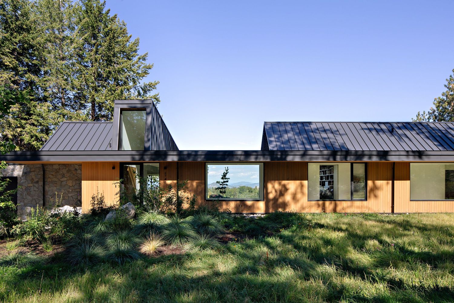 Modern wood and metal home with large windows set on grassy hillside featuring connected pavilion-style structures.
