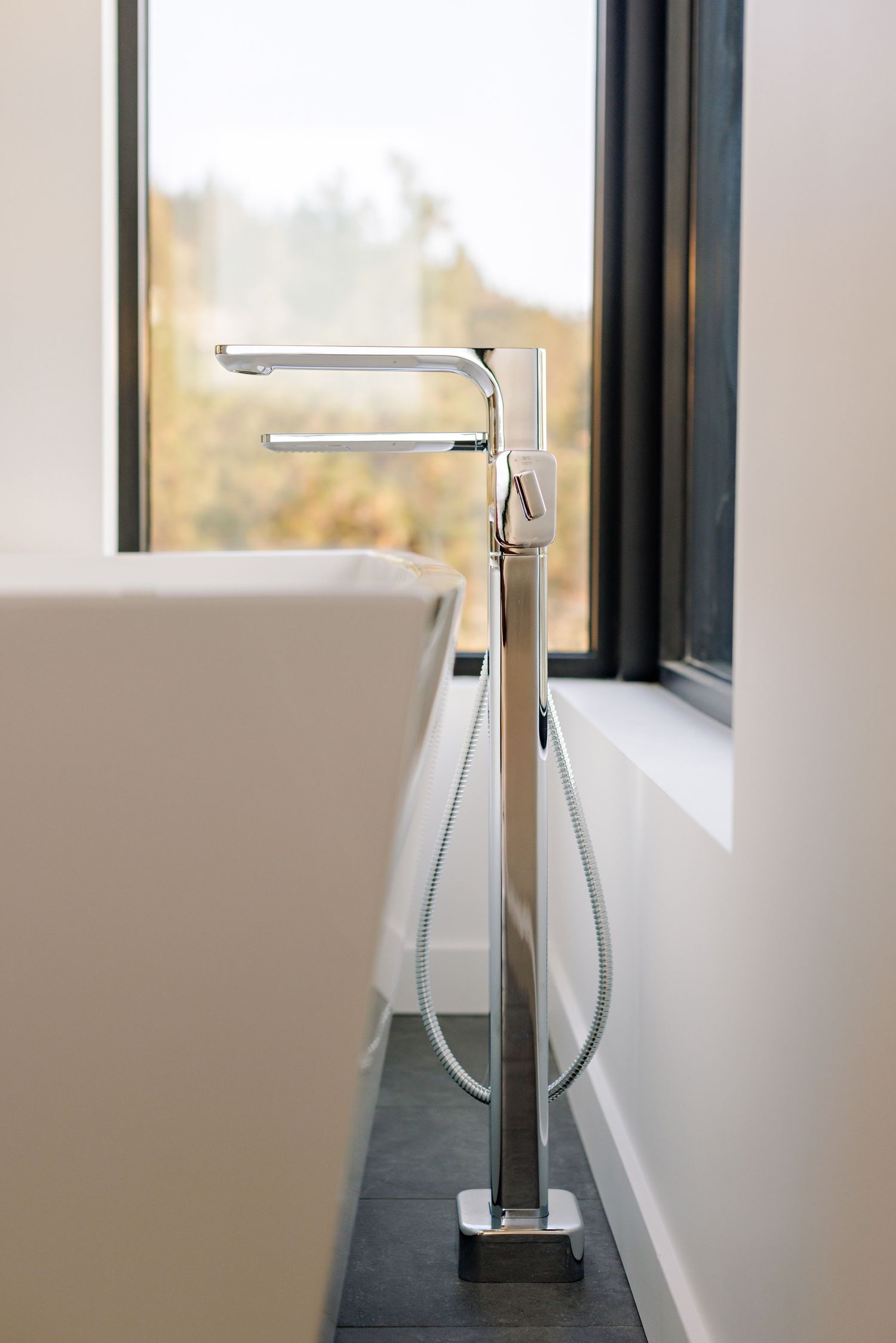 Modern freestanding bathtub faucet with handheld shower attachment mounted on dark tile floor next to white tub.