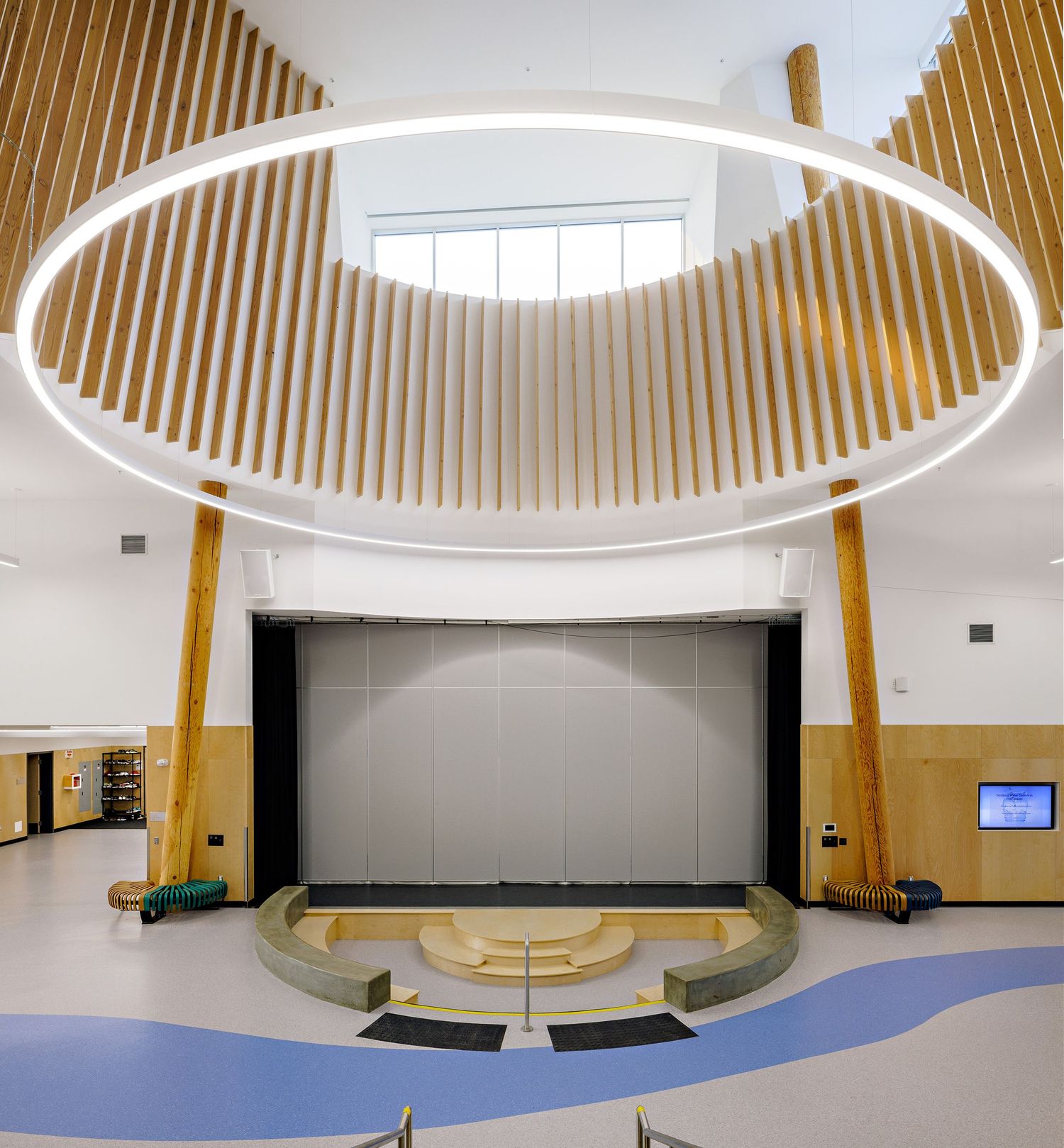 Modern auditorium with circular skylight, wooden slat walls, and curved seating facing a stage area.