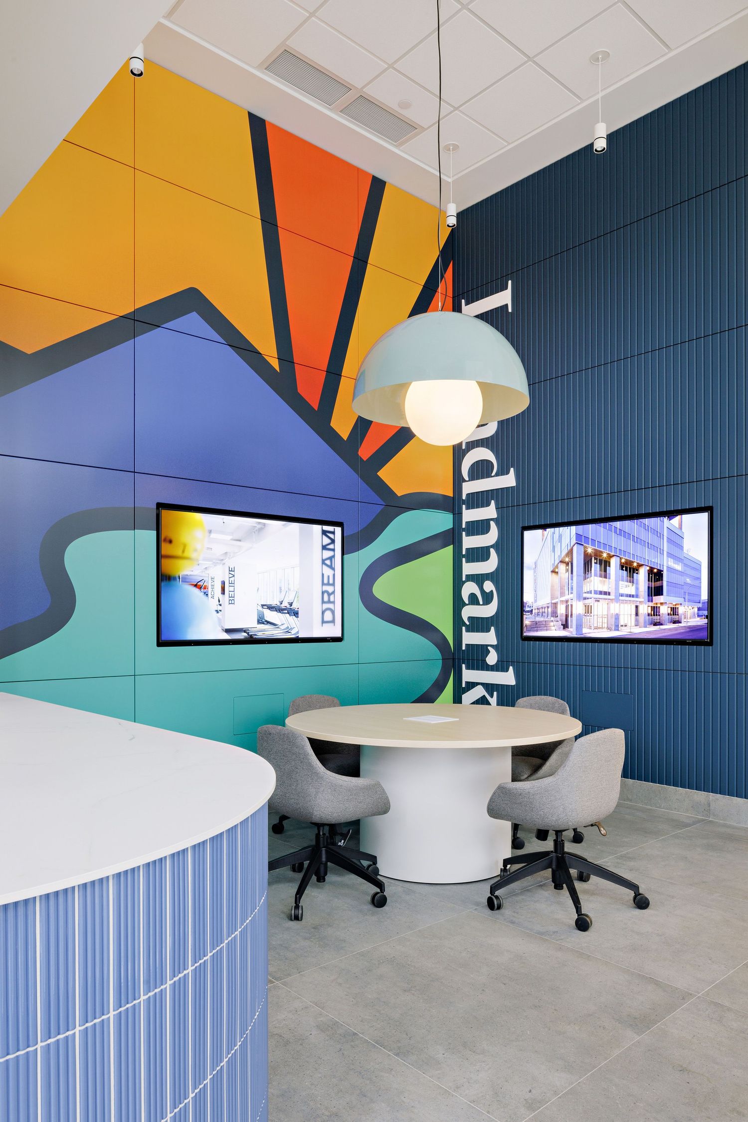 Modern office meeting area with colorful geometric wall murals, curved furniture, and mounted display screens.