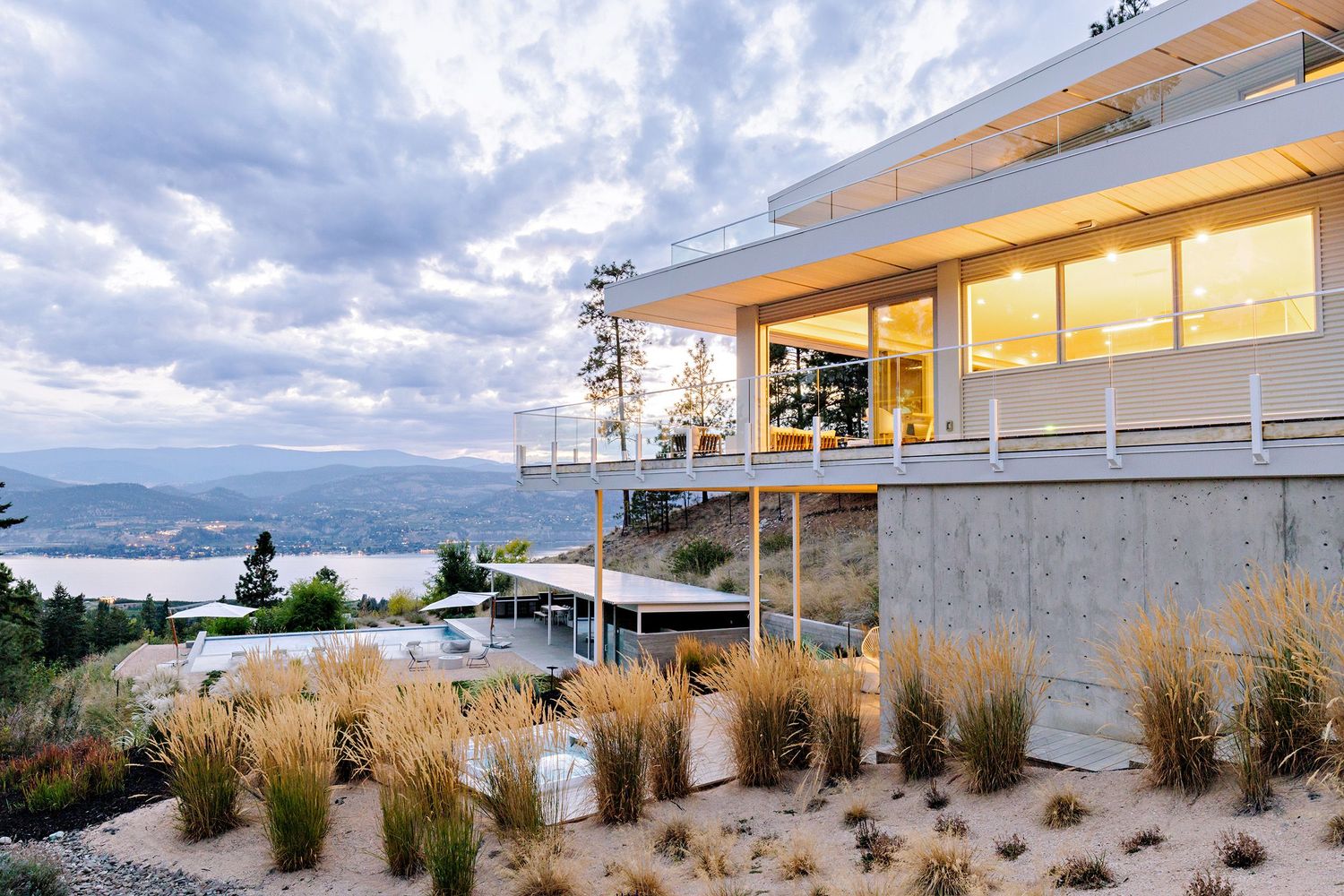 Modern hillside home with glass walls and native grasses overlooking a scenic lake under dramatic cloudy skies.