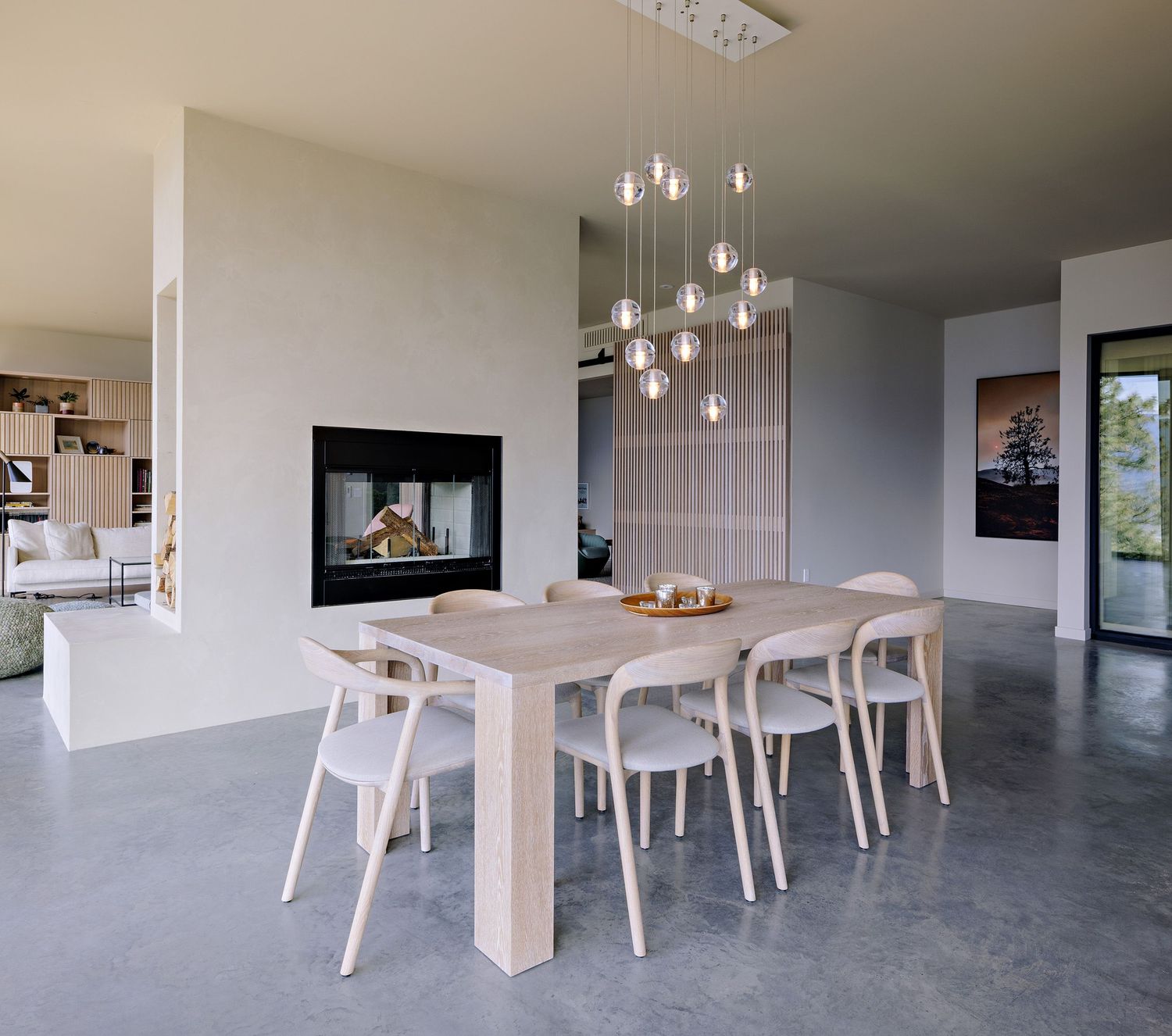 Modern dining room with light wood table, white chairs, cascading glass pendant lights and built-in fireplace on concrete floor.