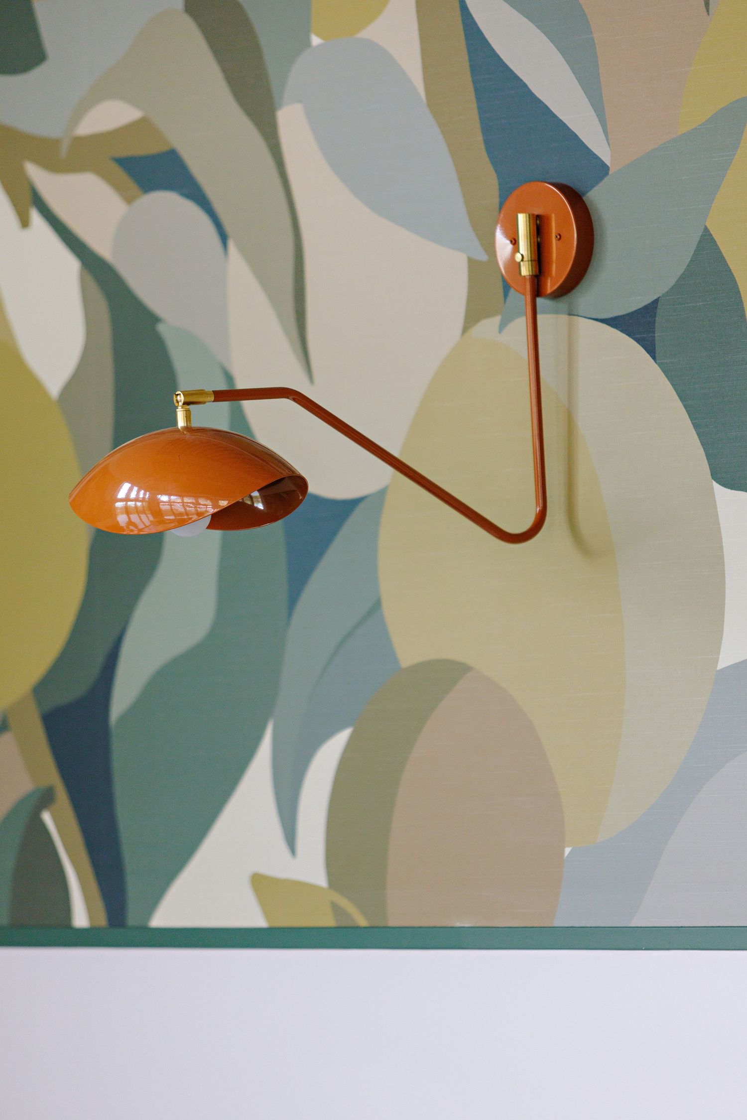 Modern orange metal wall sconce with curved arm mounted on abstract floral wallpaper in muted teal and beige tones.