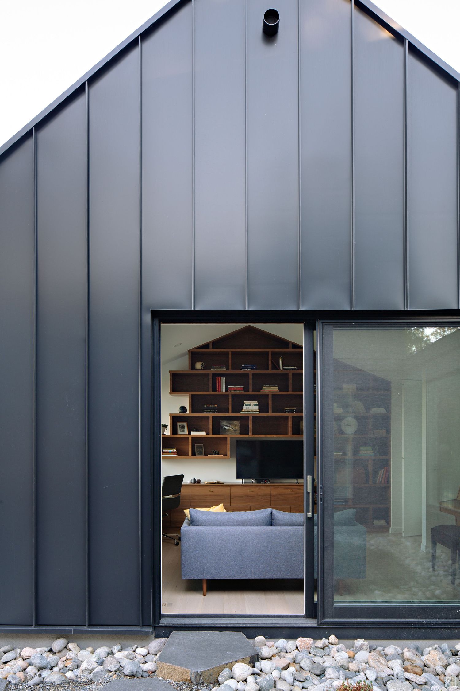 Modern gray metal-clad building exterior with sliding glass door revealing an interior office space with brick wall and shelving.