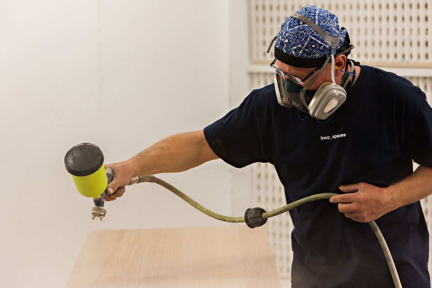 Person in protective gear uses an industrial spray gun to apply coating in a white workshop setting.