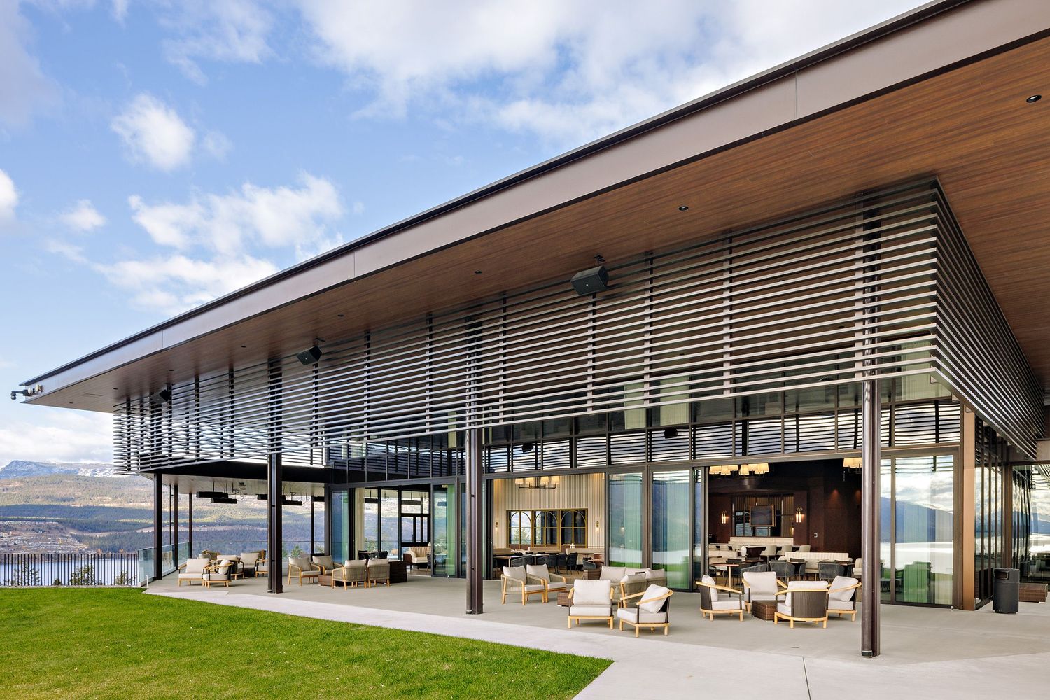 Modern luxury hillside house with glass walls, wooden overhanging roof and outdoor seating overlooking scenic landscape.