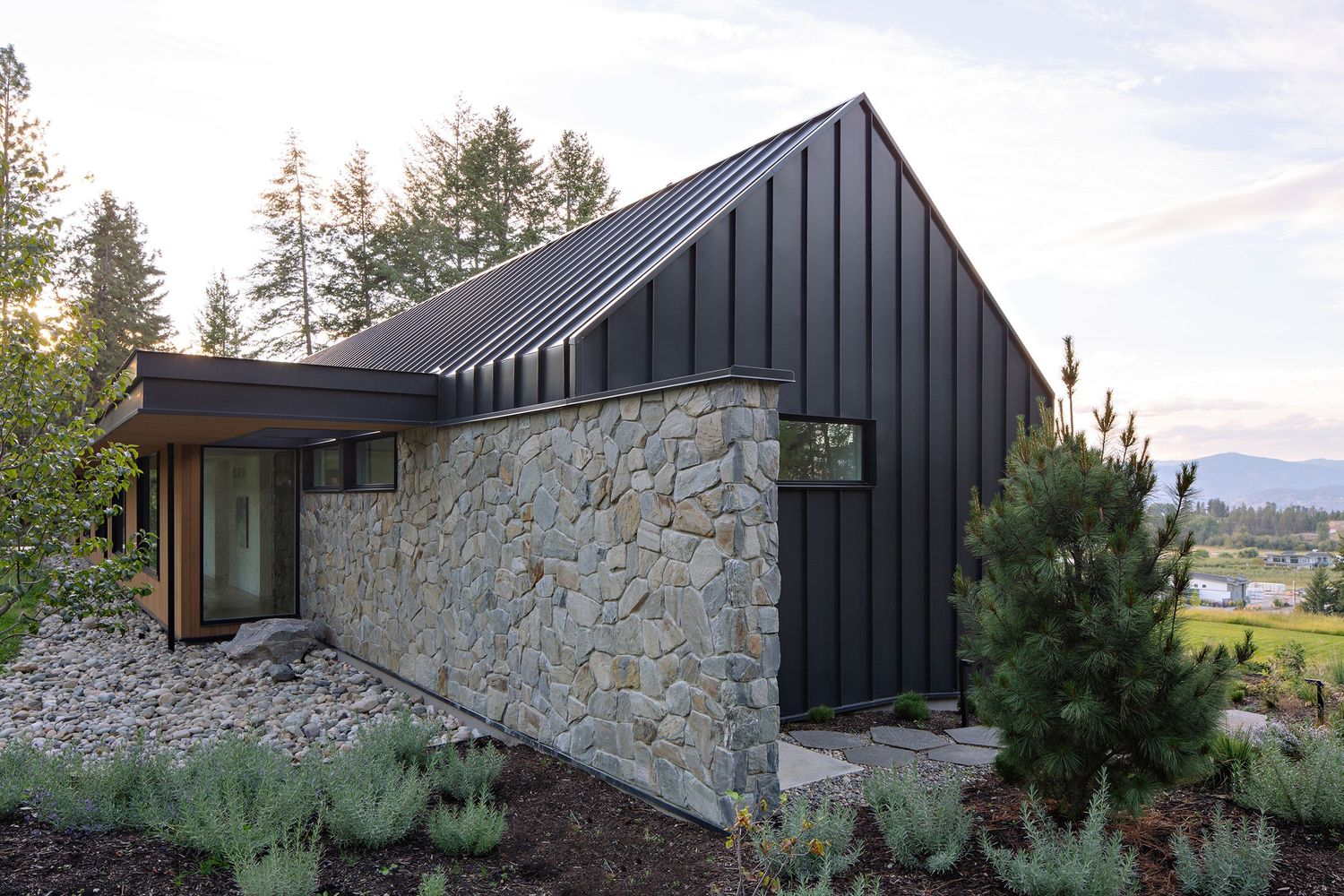 Modern mountain home featuring dark metal cladding, stone walls, and angled rooflines against a wooded landscape.