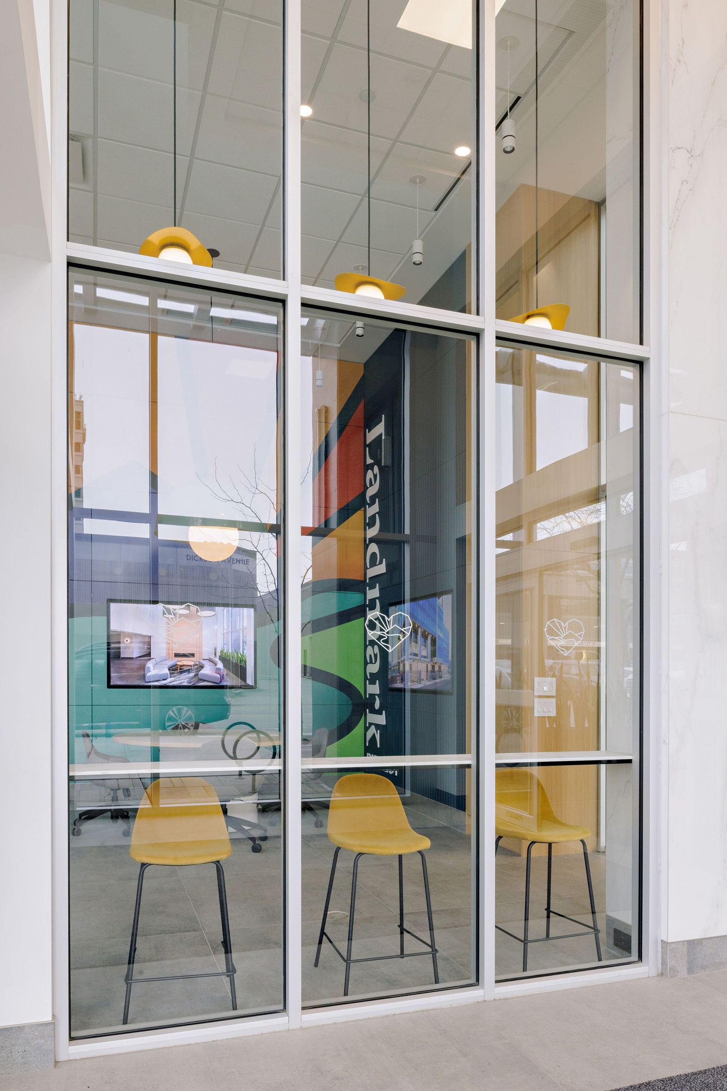 Modern office interior with yellow bar stools visible through glass windows, featuring colorful wall art and pendant lighting.