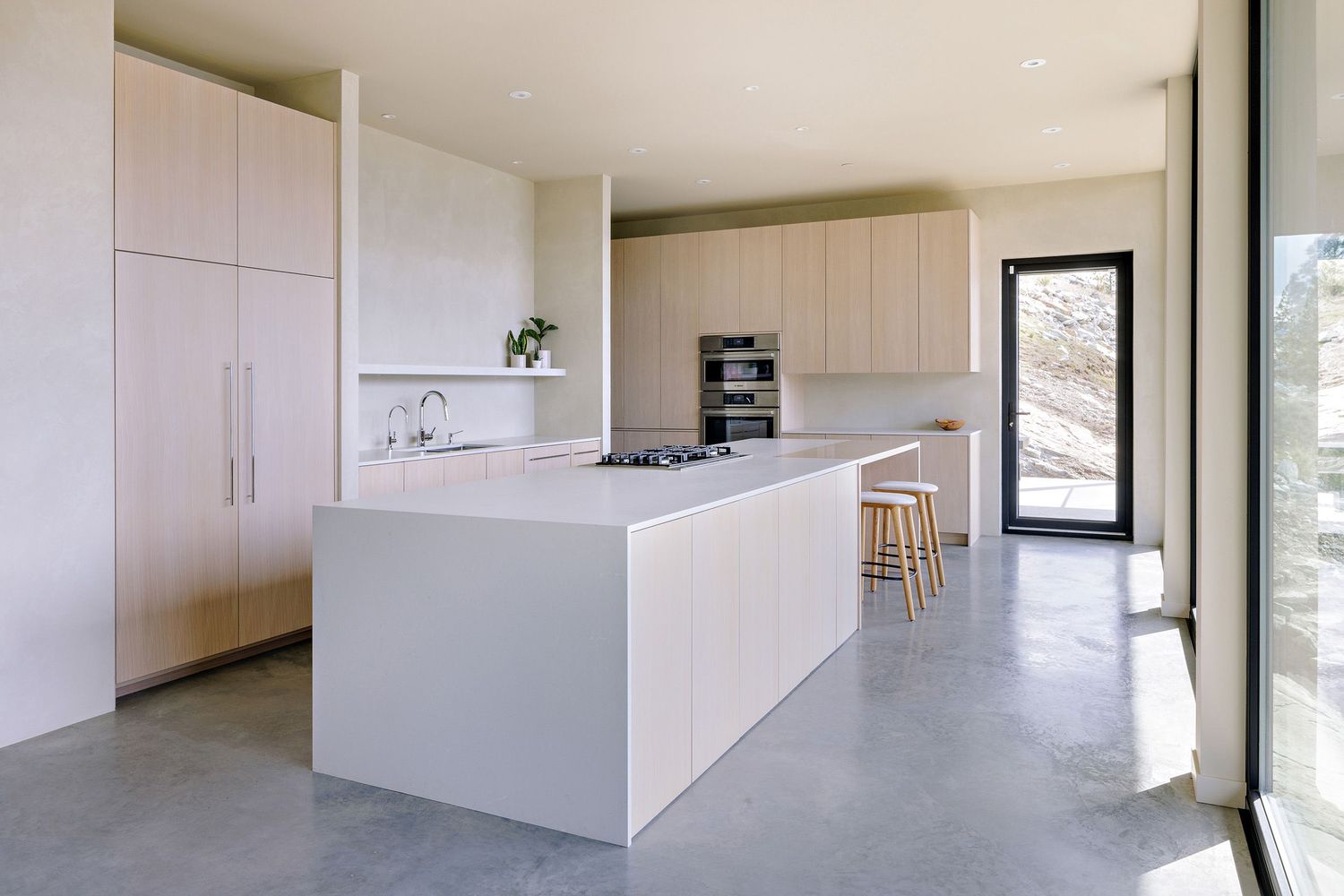 Modern minimalist white kitchen with sleek island, concrete floors, and floor-to-ceiling cabinets in a bright space.