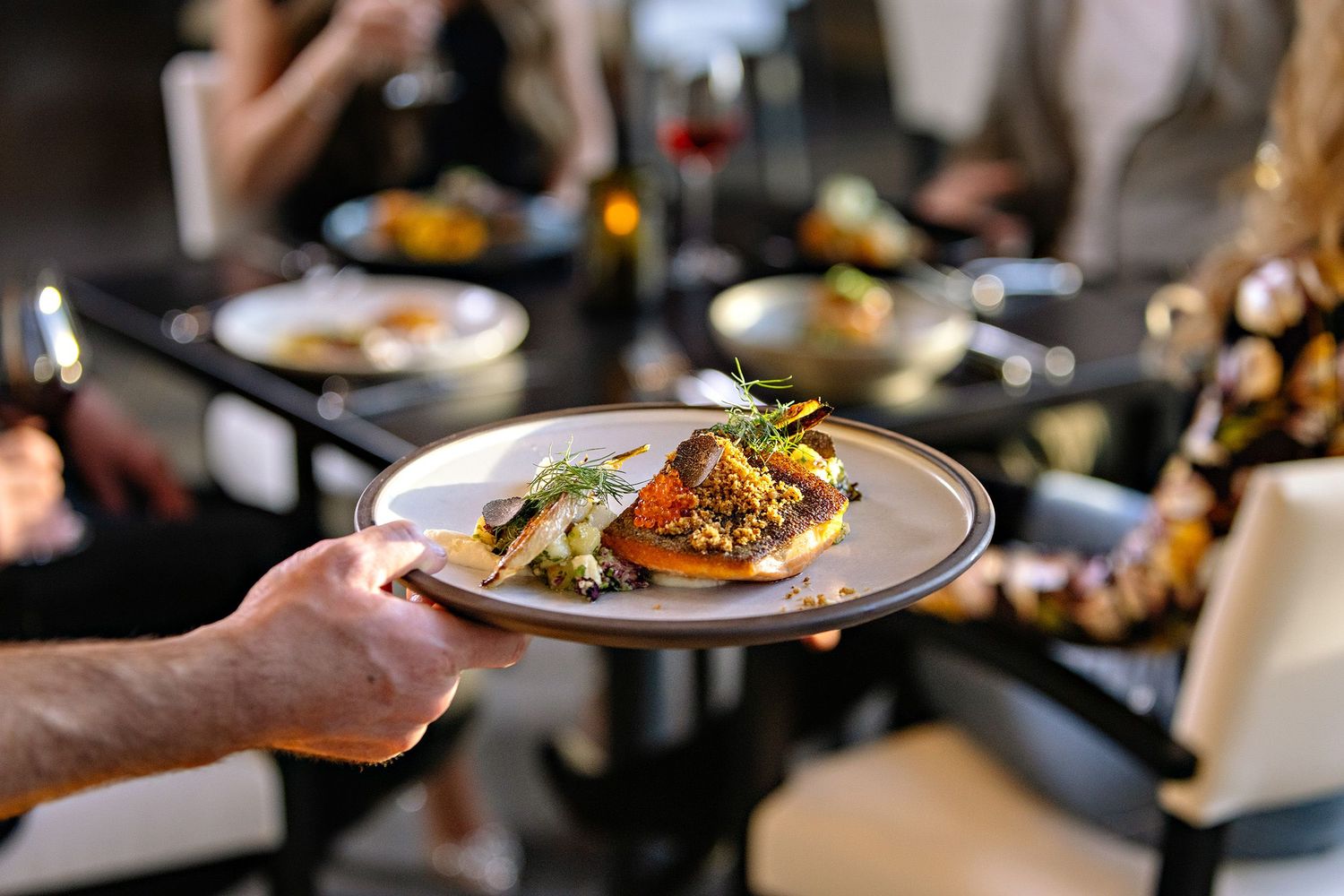 Server carrying a beautifully plated dish with garnished seafood at an upscale restaurant during dinner service.