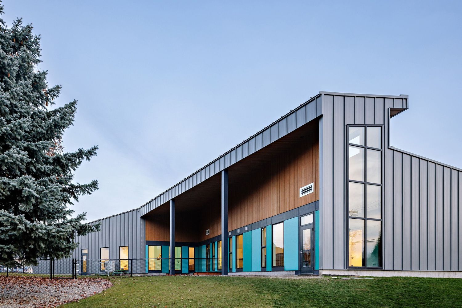 Modern angular building with turquoise windows and vertical metal siding set against evergreen trees and cloudy sky.