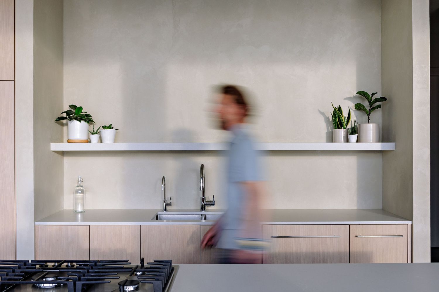Motion blur of someone walking past a modern minimalist kitchen with floating shelves and potted plants.