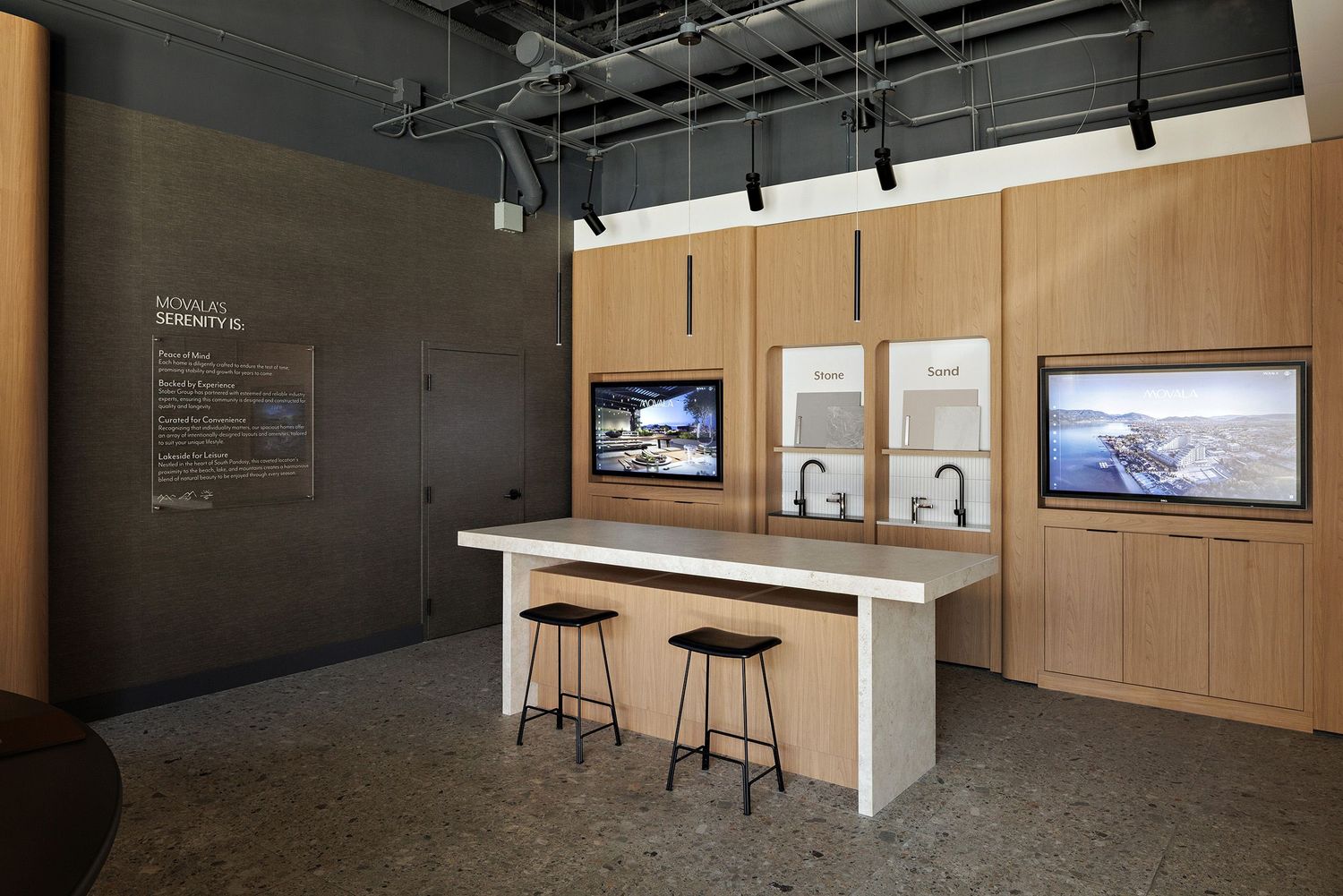 Modern office space with wooden cabinets, white counter, bar stools, and mounted displays on a dark wall.