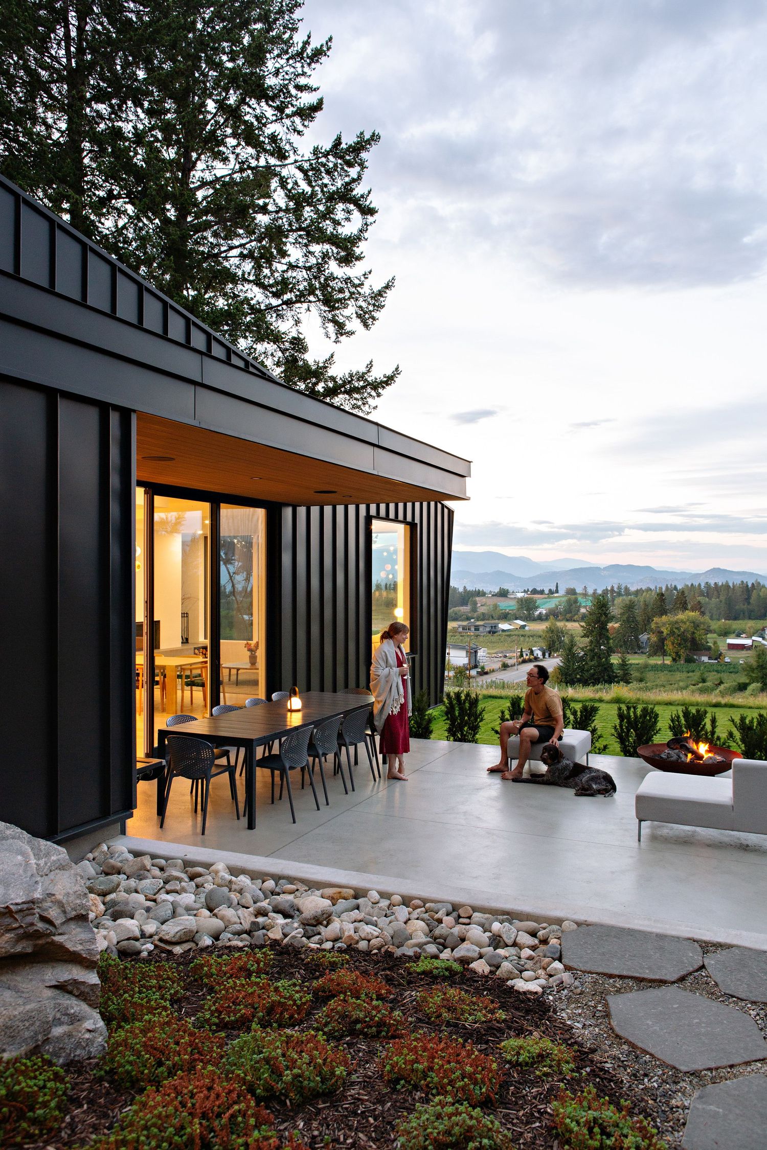 Modern black container home with outdoor patio and dining area overlooking scenic mountain landscape at dusk.