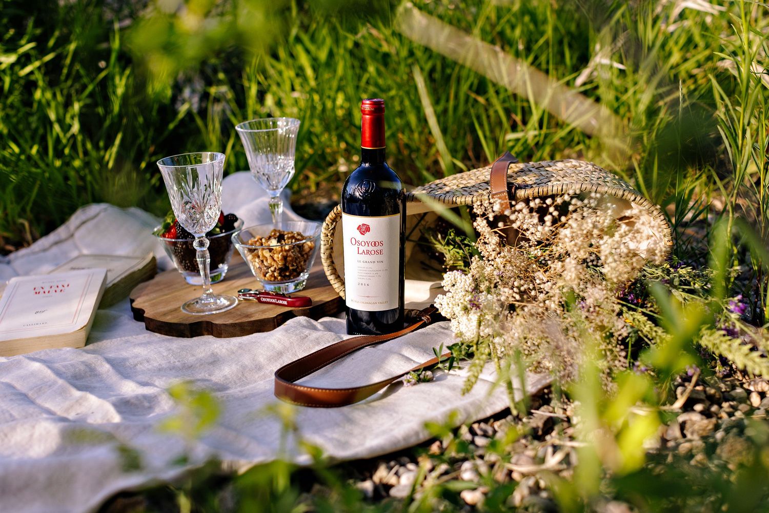 A romantic outdoor picnic setup with wine bottle, glasses, and delicate white flowers on a blanket in lush grass.