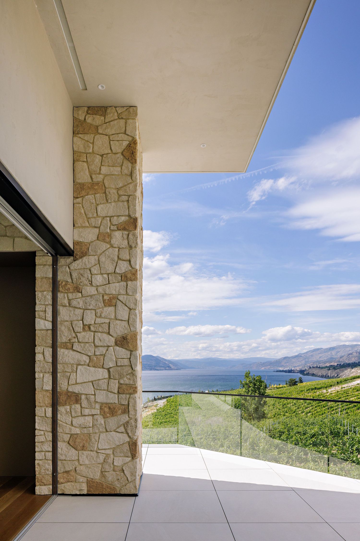 Stone pillar and glass railing on a modern patio with panoramic views of a lake and rolling vineyards.