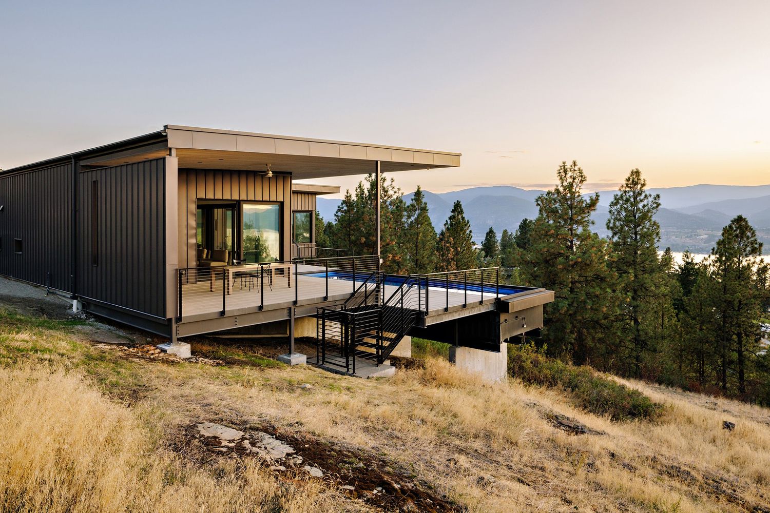 Modern hillside home with cantilevered deck overlooking mountain valley and lake views at sunset.