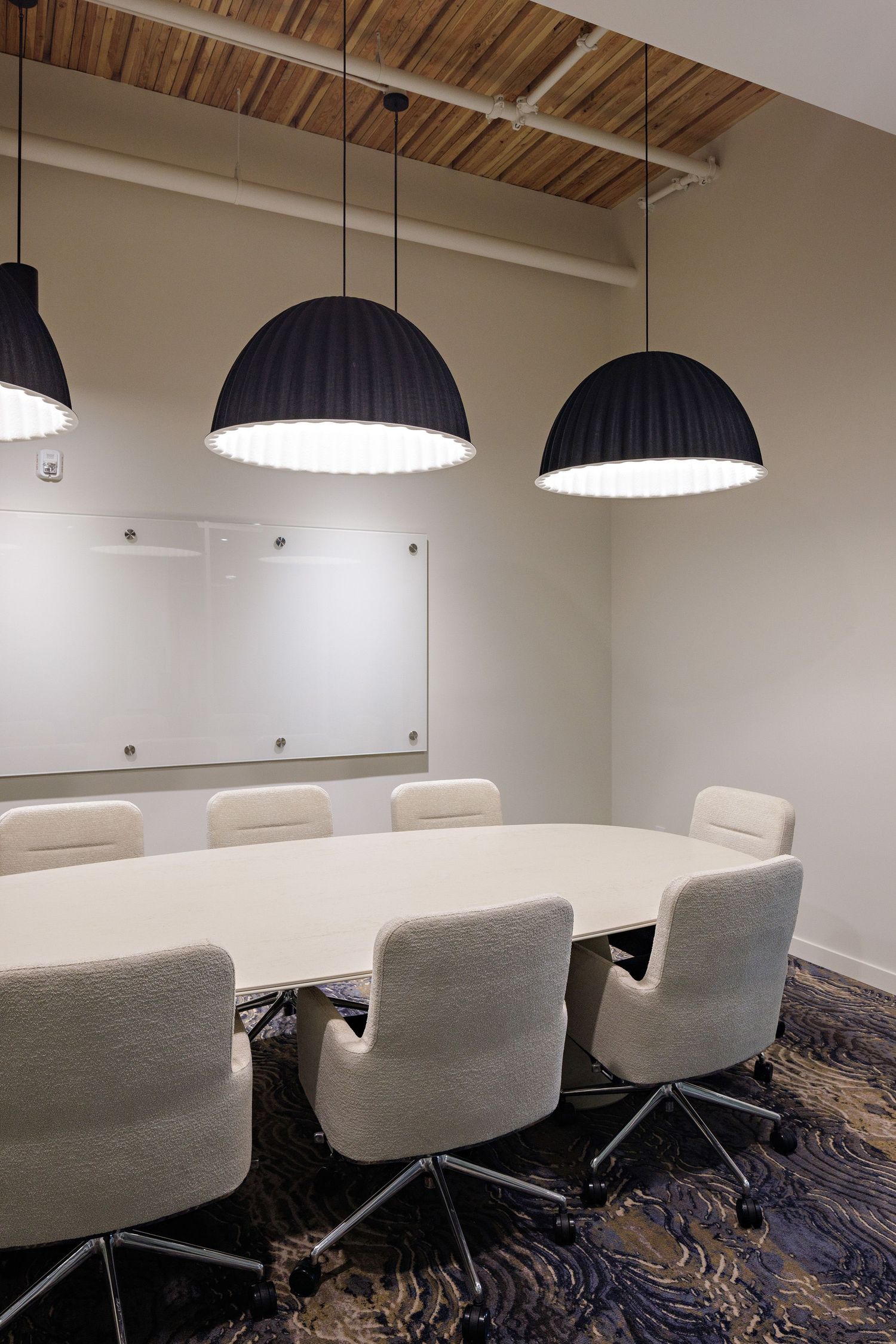 Modern conference room with white oval table, gray upholstered chairs and three large black dome pendant lights hanging above.