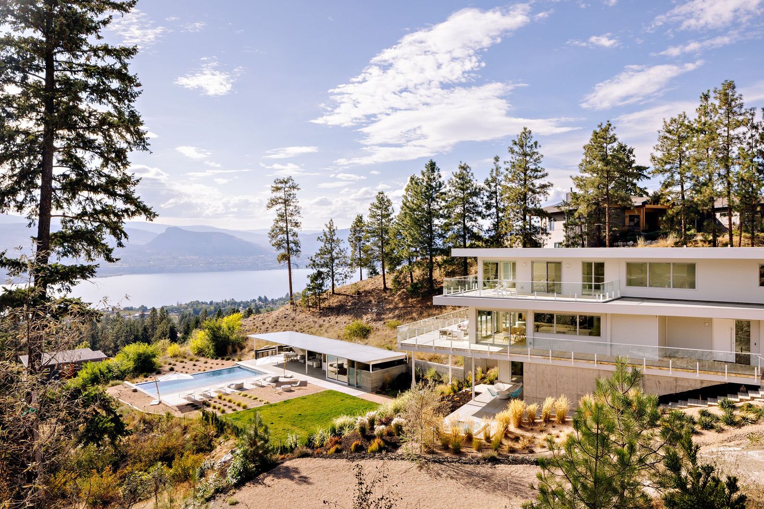 Modern luxury hillside home with infinity pool overlooking mountains and lake through tall pine trees.