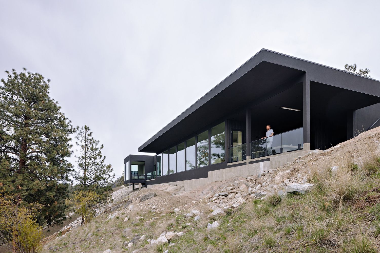 A modern black hillside house with floor-to-ceiling windows perched on a rocky slope among trees and natural terrain.