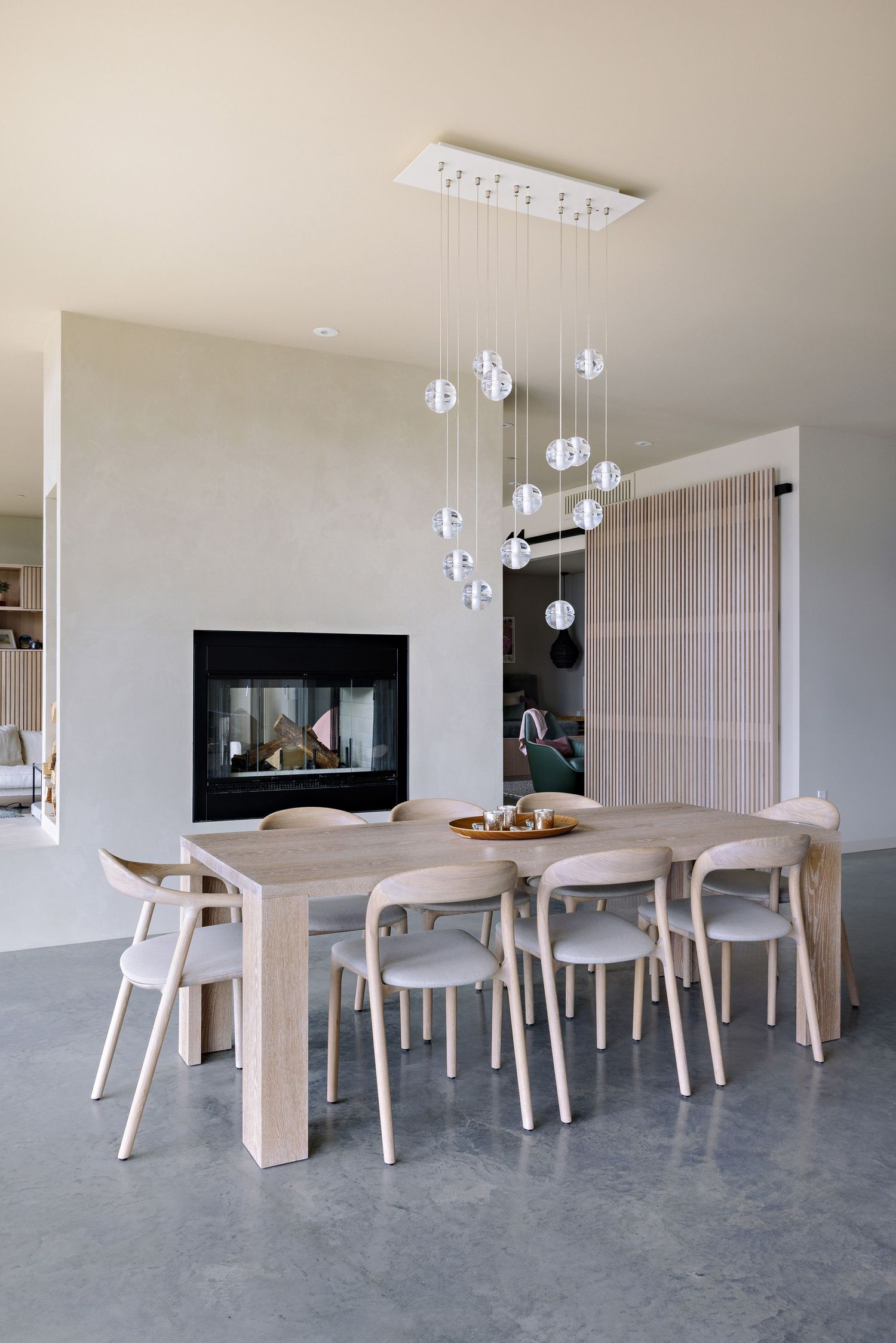 Modern dining room with hanging glass pendant lights, wooden table, and built-in fireplace on concrete floors.