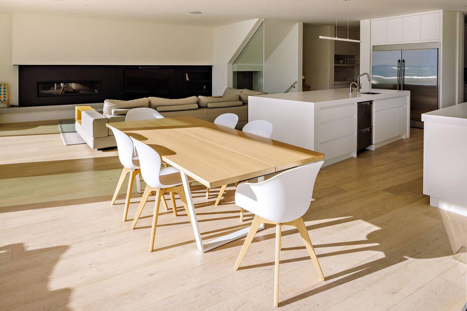 Modern open concept dining space with wooden table, white chairs and natural light casting geometric shadows on hardwood floors.