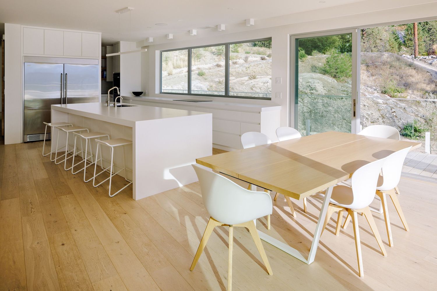 Modern open concept kitchen and dining area with white island, bar stools, and wooden table in bright minimalist space.