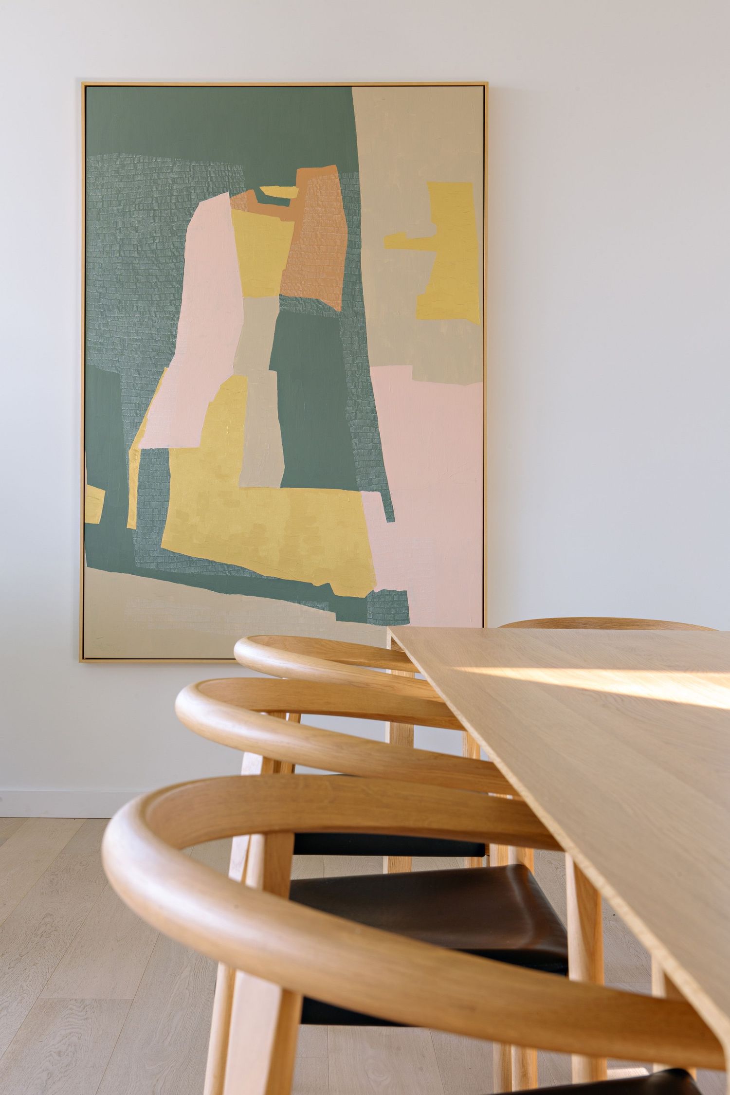 Modern abstract art print with muted pink, yellow and sage green shapes hangs above wooden dining chairs in minimalist room.