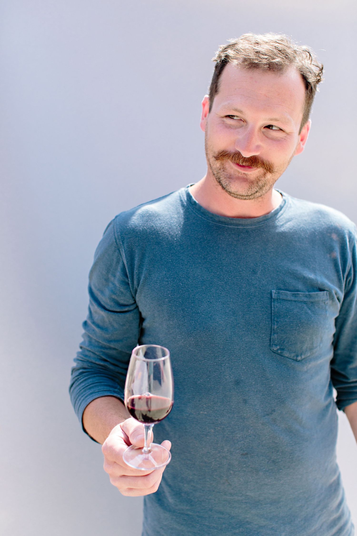 Person in blue sweater smiling while holding a glass of red wine against light background.