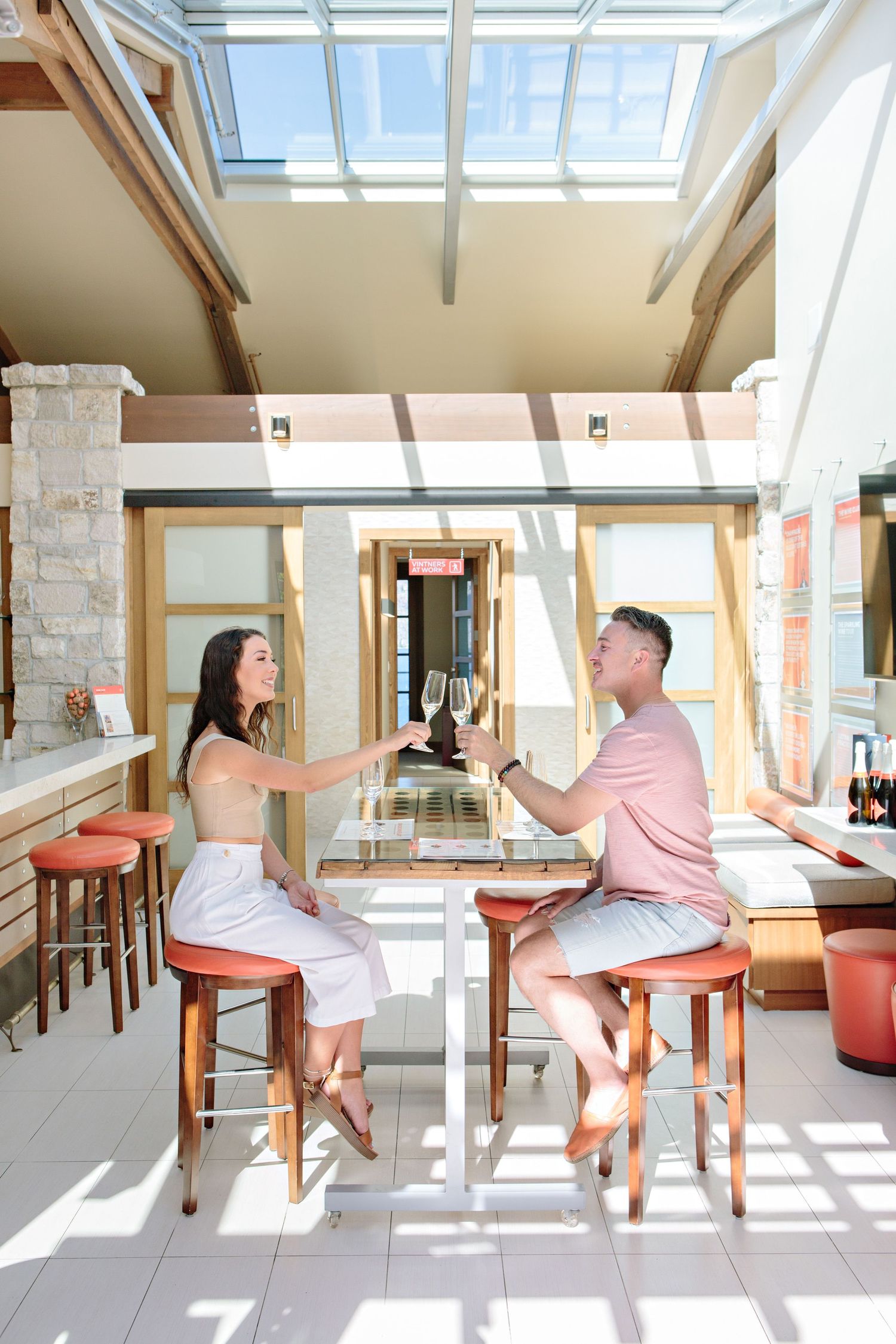 Couple toasting drinks at sunlit bar with skylight and rustic decor.