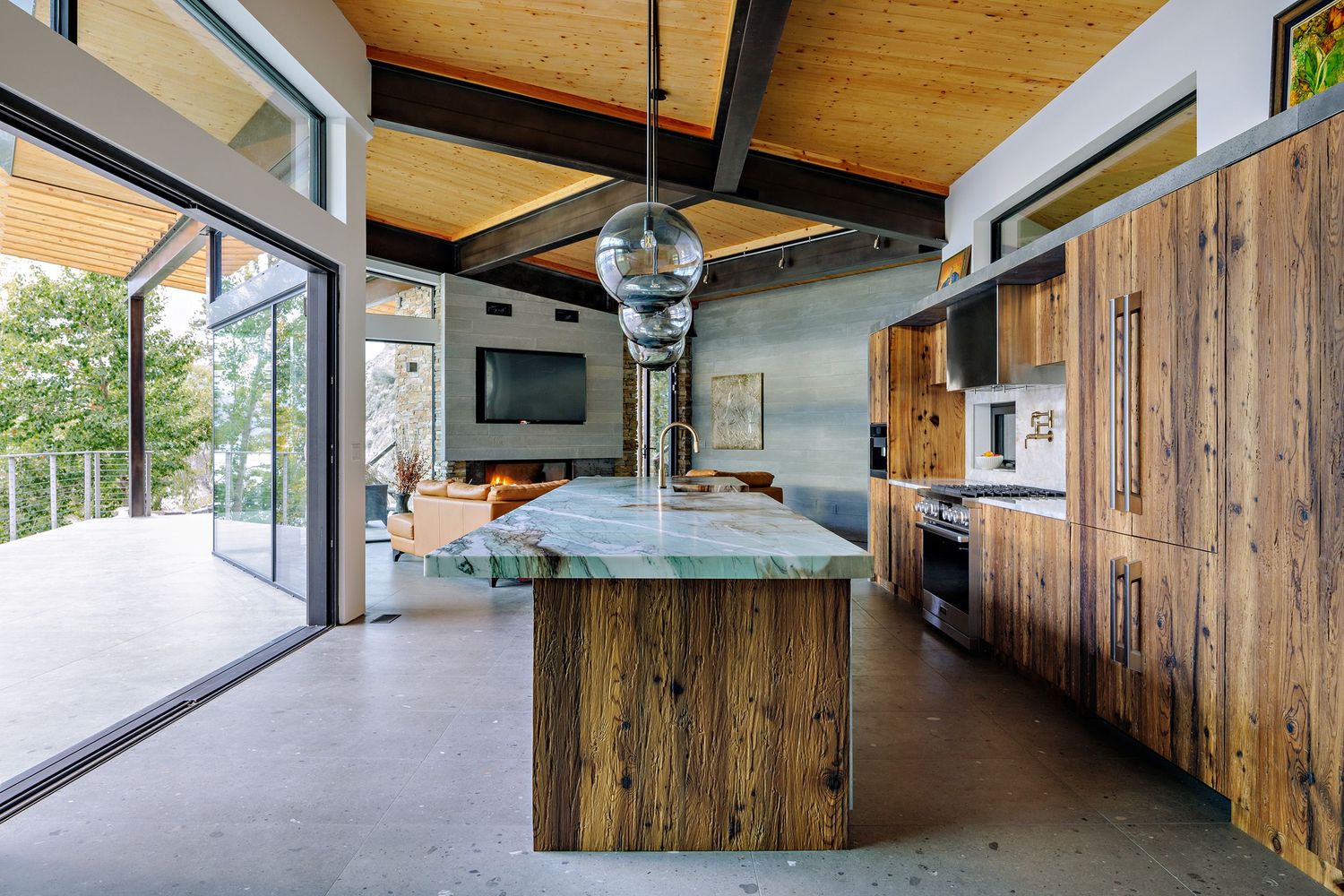 Modern kitchen with rustic wood cabinets, concrete floors, and floor-to-ceiling windows overlooking nature.